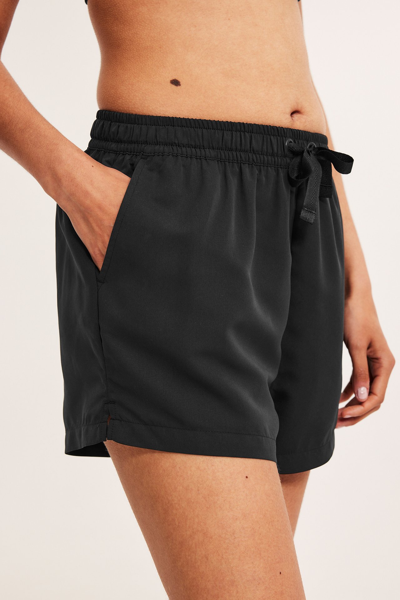 Sidebilde av en kvinne i svarte vevde badeshorts med snøring i midjen og lommer, hånd i lommen.