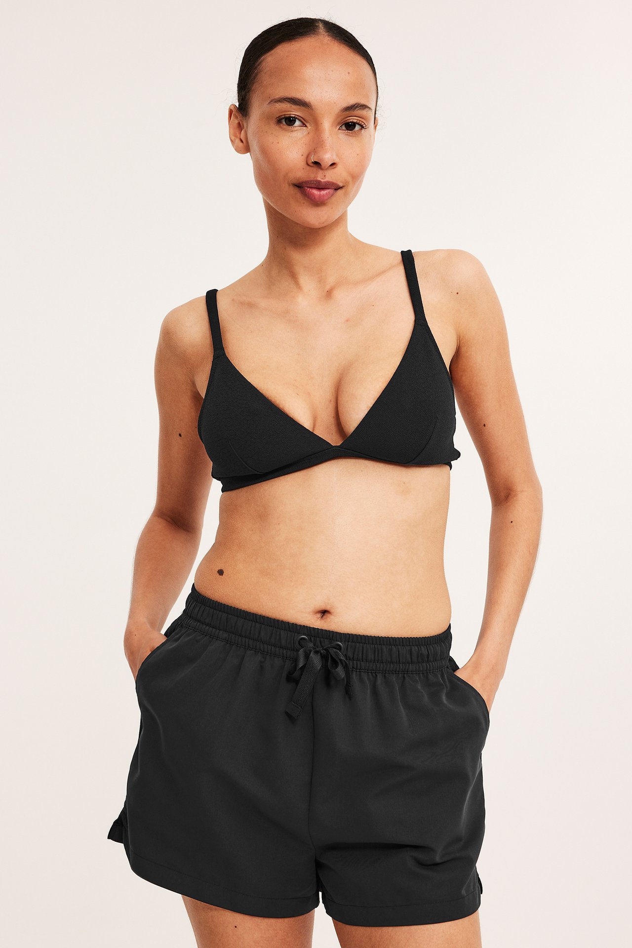 Frontbilde av kvinne i svarte vevde badeshorts med snøring og en svart bikinitopp.