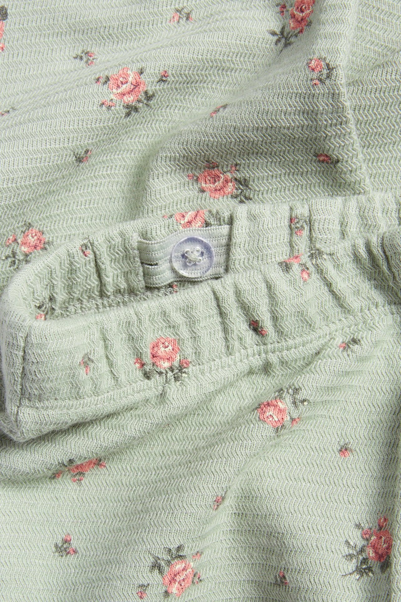 Zbliżenie zielonych, teksturowanych, rozszerzanych legginsów w drobny różowy kwiatowy wzór z regulowanym pasem na guzik.