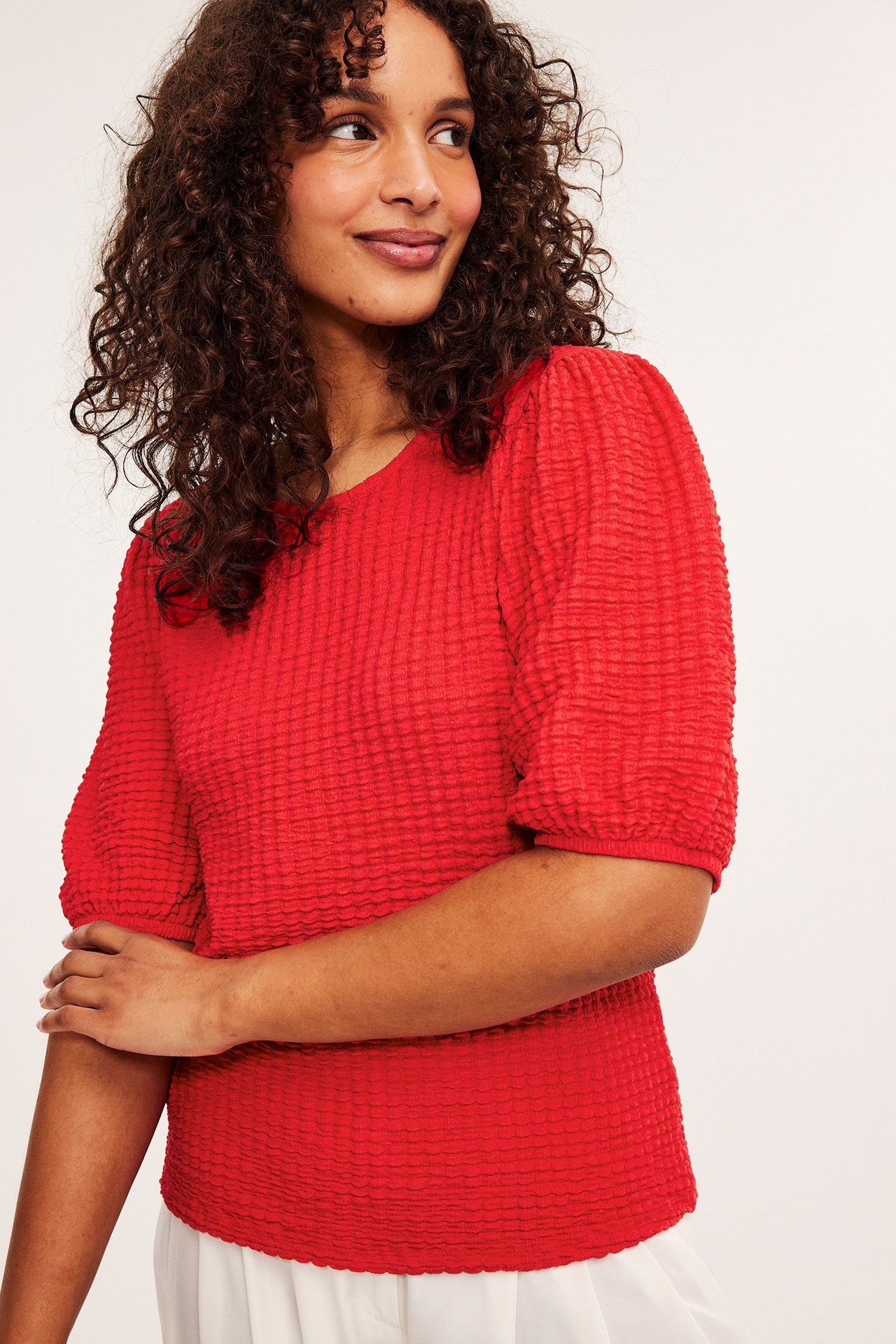 Forfra: kvinne i rød strukturert topp med puffermer og hvite bukser.