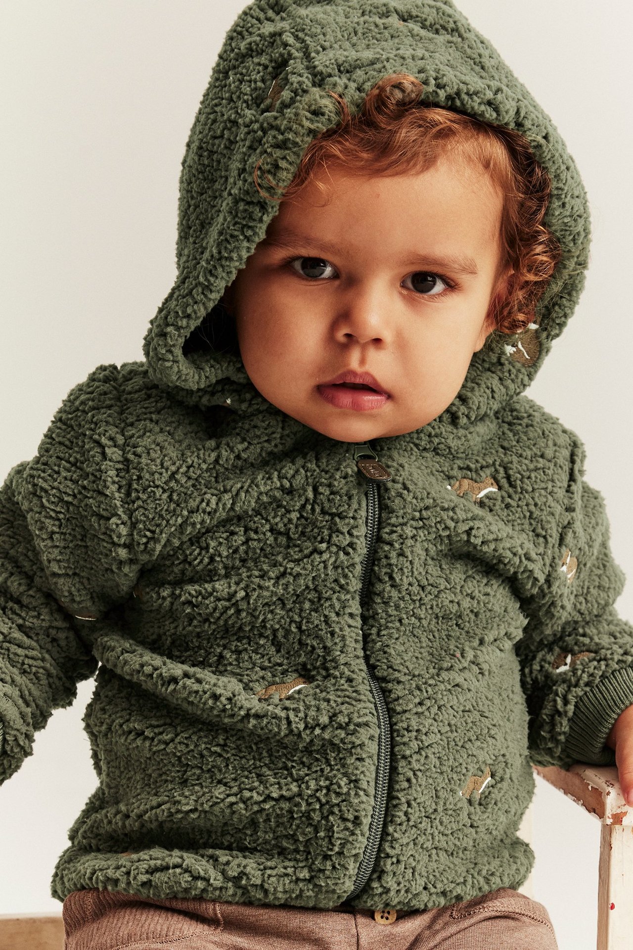 Närbild framifrån på en baby i en mörkgrön pilejacka med huva, rävbrodyr och dragkedja.