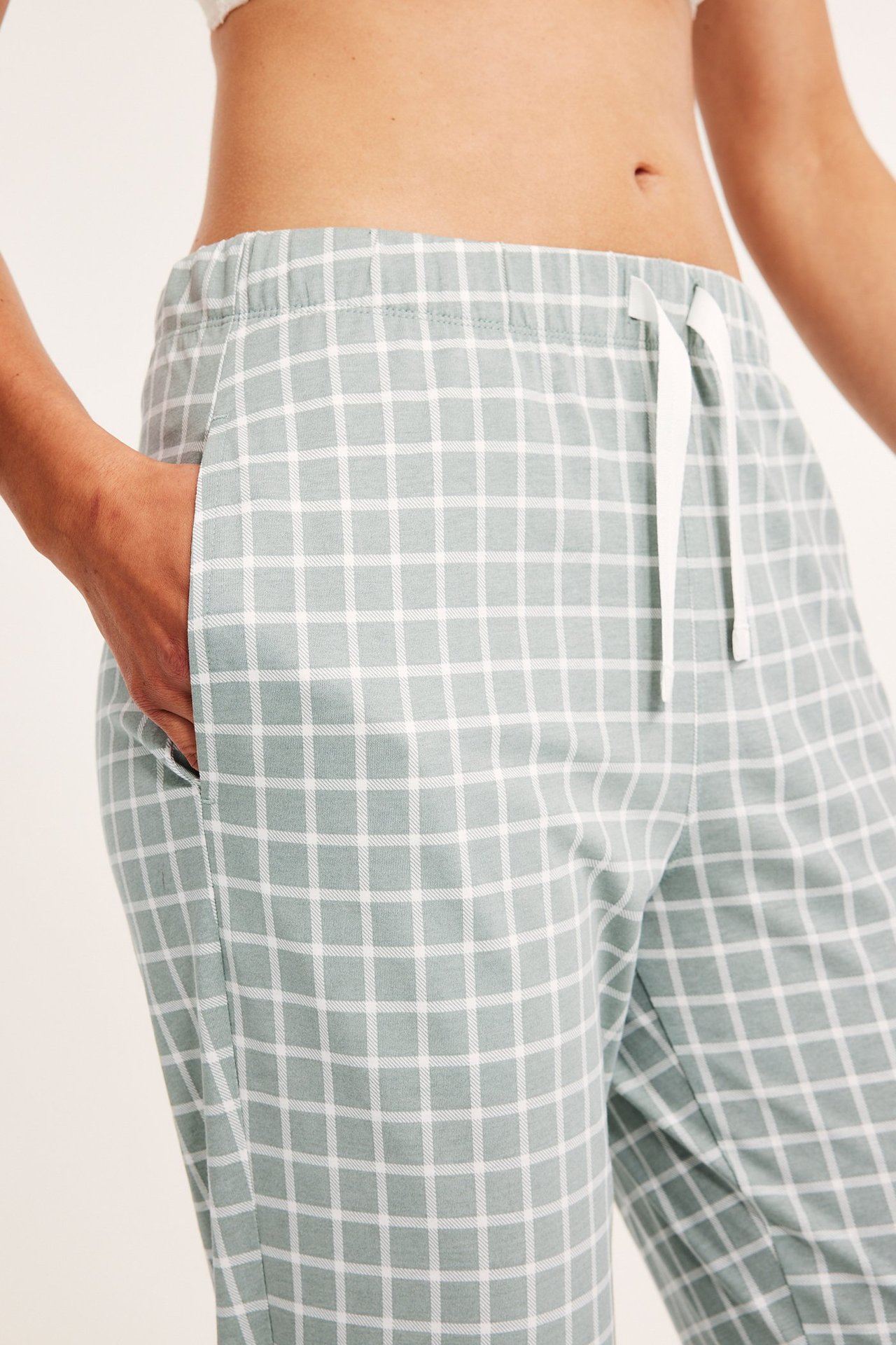 Lähikuva naisesta yllään vaaleanvihreät ruudulliset puuvillapyjamahousut joustovyötäröllä, käsi taskussa.