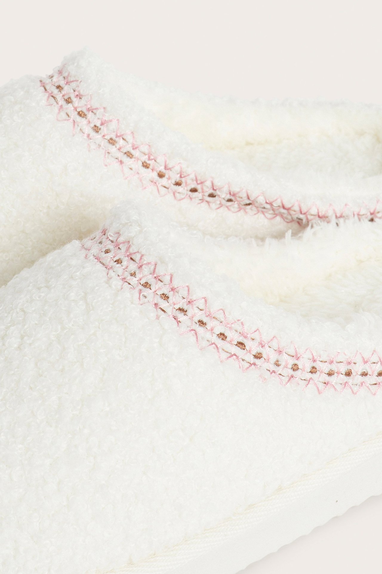 Zbliżenie na kremowe kapcie z puszystego materiału z różowo-brązowym haftowanym brzegiem.