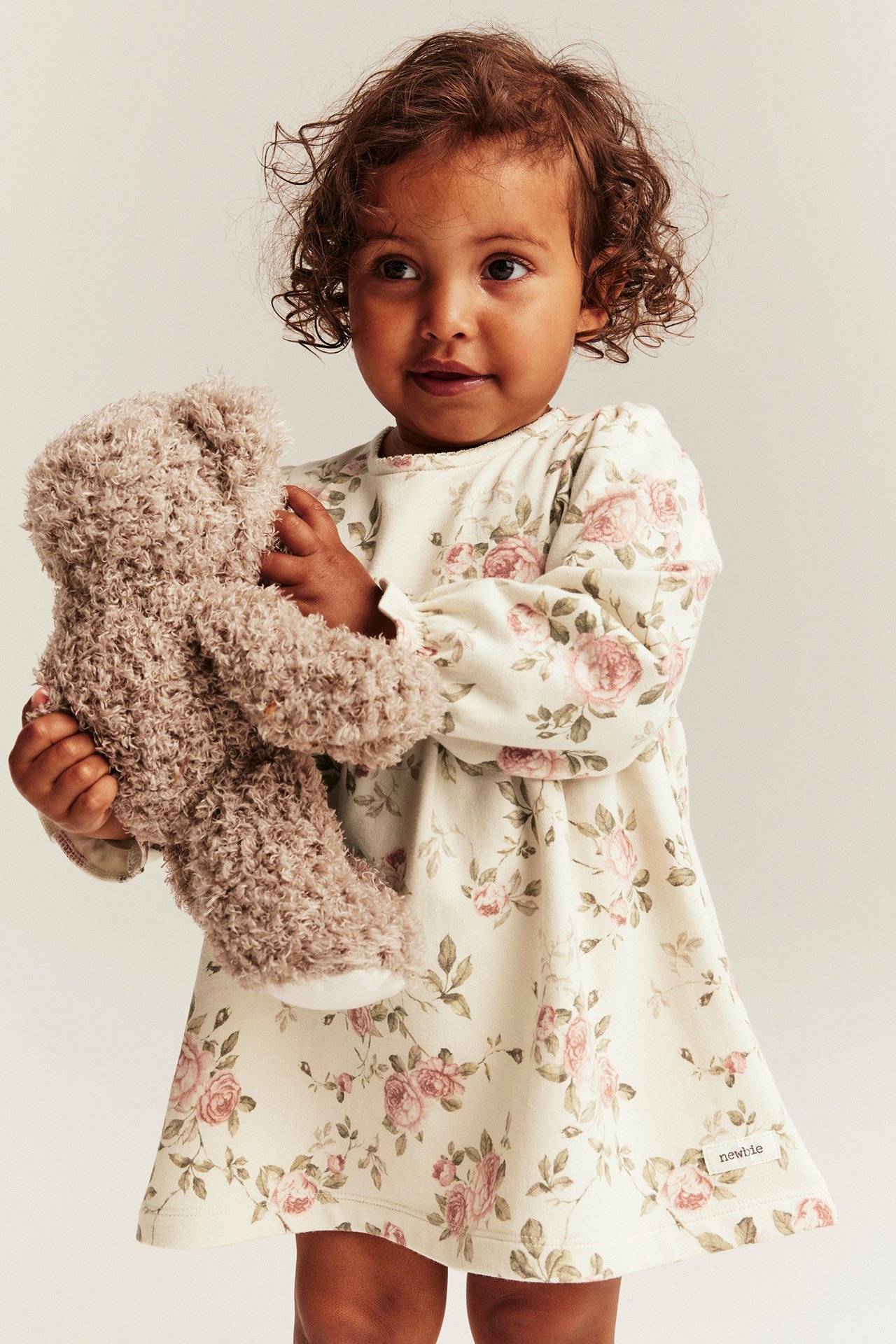 Forfra: Et barn i lys beige blomstret jerseykjole med puffermer, holder en brun teddybjørn.