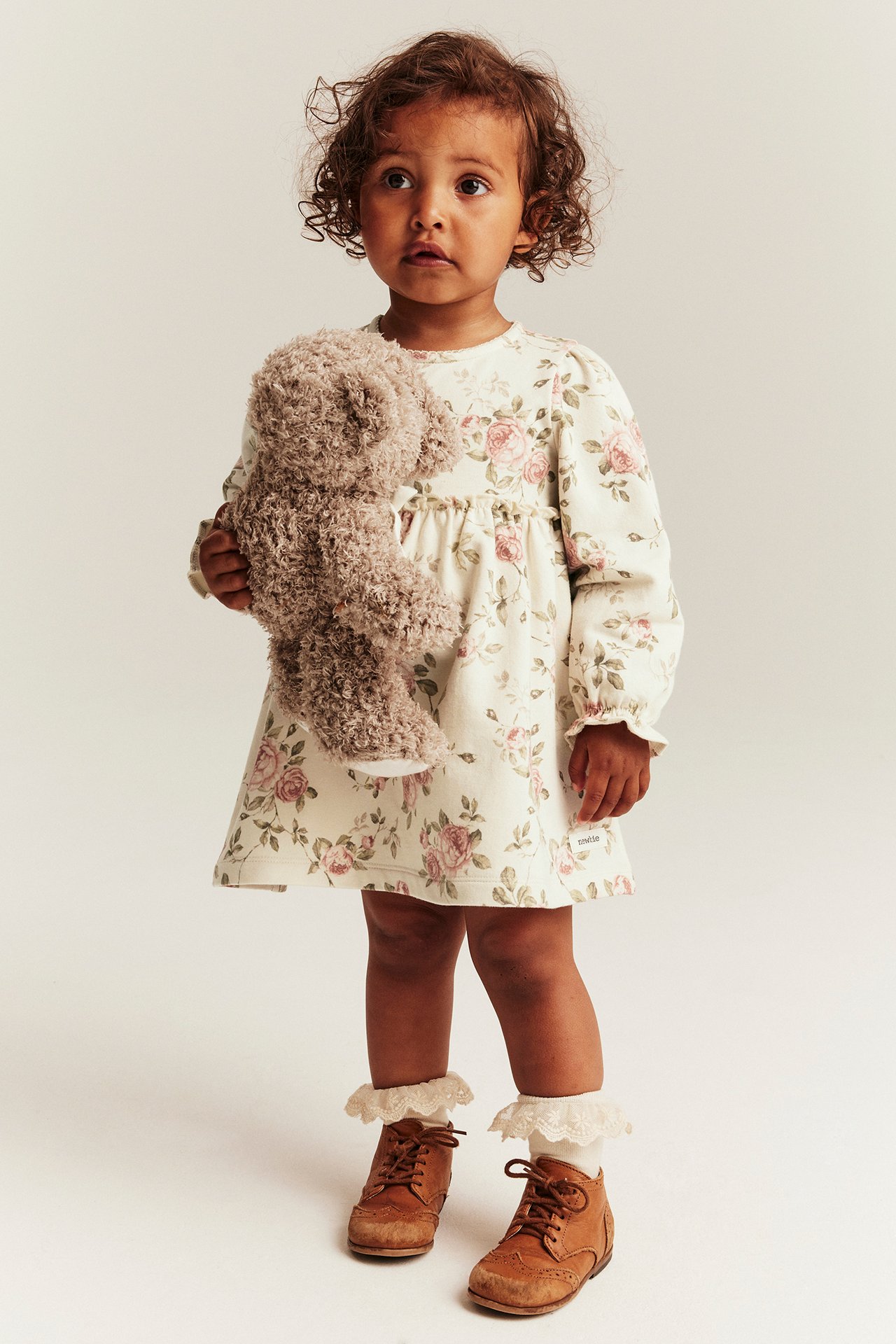 Widok z przodu: Dziecko w jasnobeżowej kwiecistej sukience dżersejowej, trzyma brązowego misia, białe koronkowe skarpetki, brązowe buty.