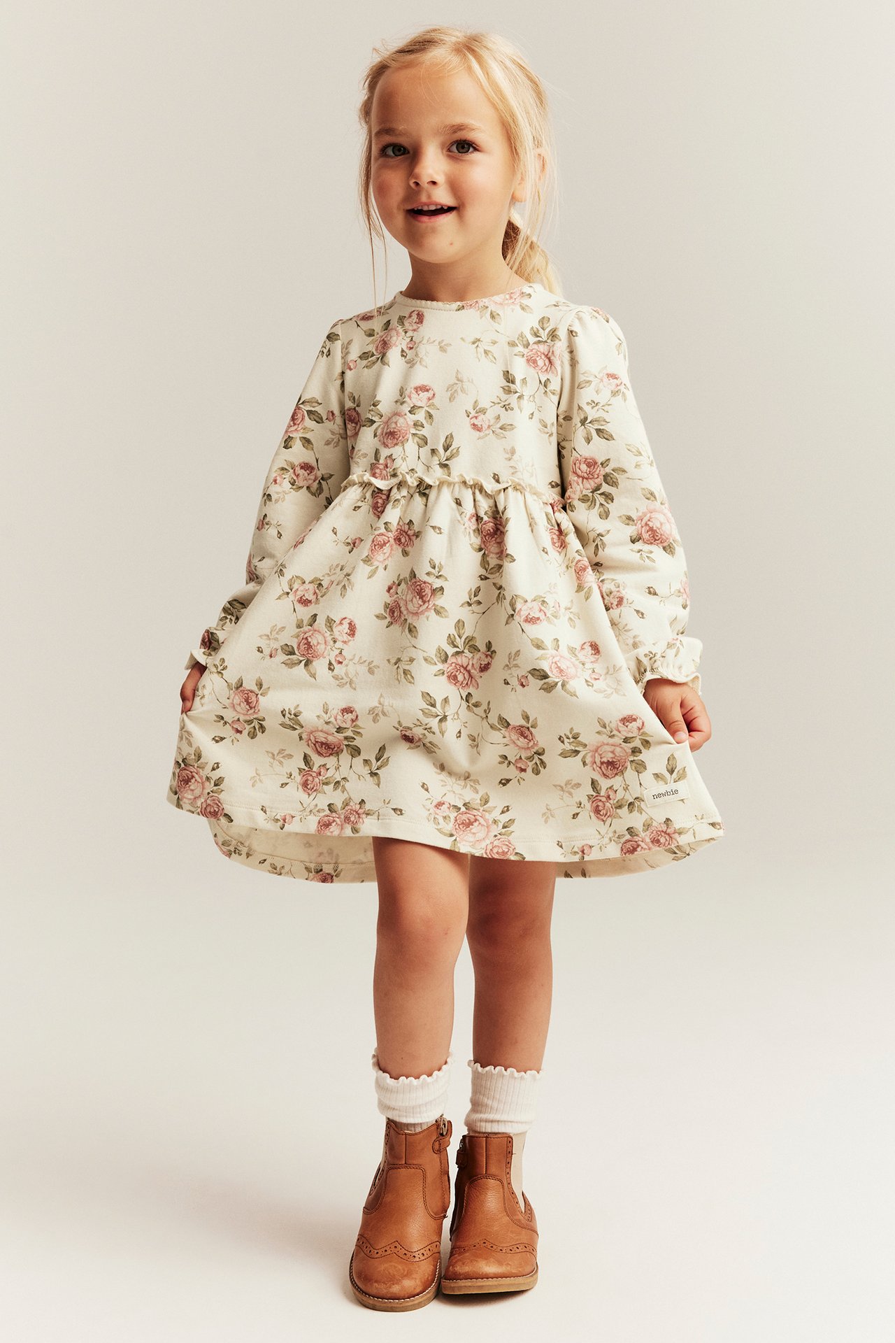 Forfra: en jente i lys beige blomstret jerseykjole med puffermer, hvite sokker, brune skinnstøvler.