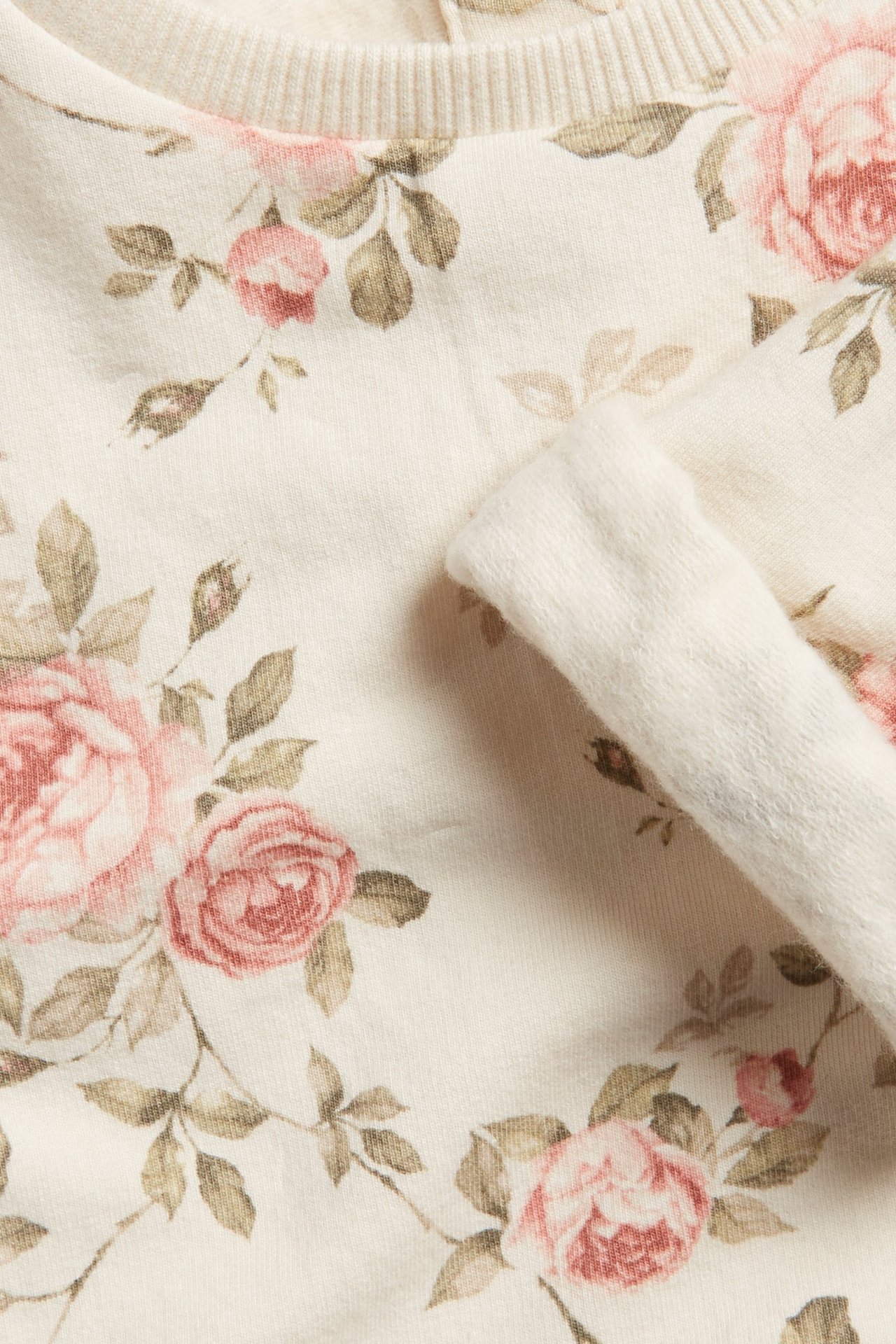 Zbliżenie na jasnobeżową bluzę w kwiaty z różowymi różami i zielonymi liśćmi, widoczny prążkowany kołnierz.
