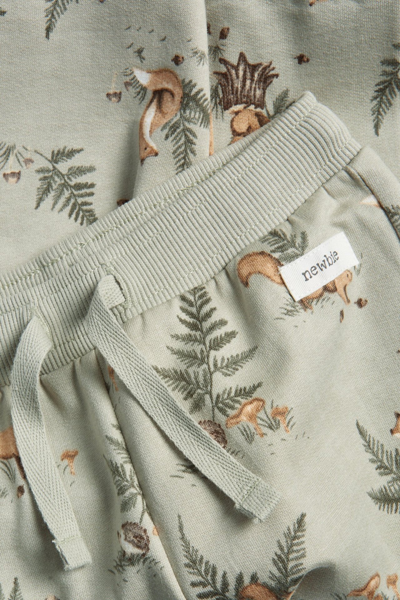 Nærbilde av lysegrønne bomullsjoggebukser med skogsmotiv, ribbet elastisk linning og snøring.