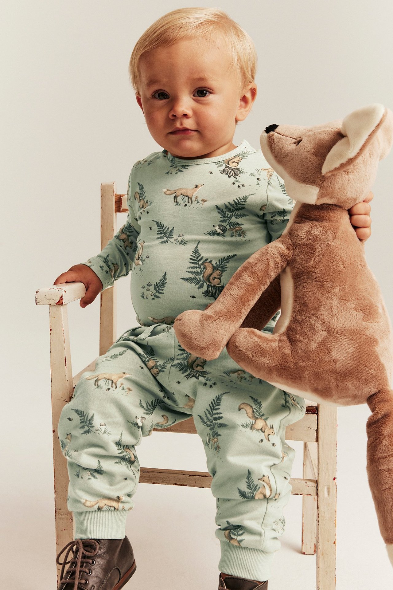 Frontalvisning: Baby i lysegrønne joggebukser med skogsmønster, matchende topp, brune sko, holder reveleke.