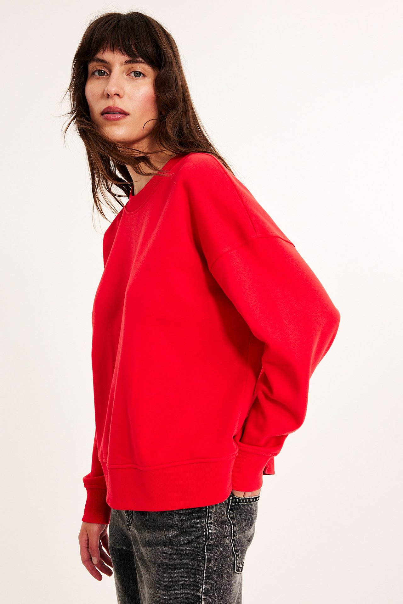 Widok z boku kobiety w czerwonej bluzie z okrągłym dekoltem, długimi rękawami i ciemnoszarych dżinsach.