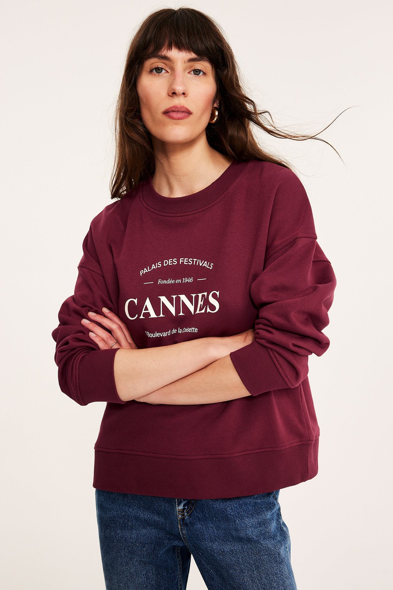 Forfra: kvinne i vinrød sweatshirt med hvitt "Cannes"-trykk og blå jeans.