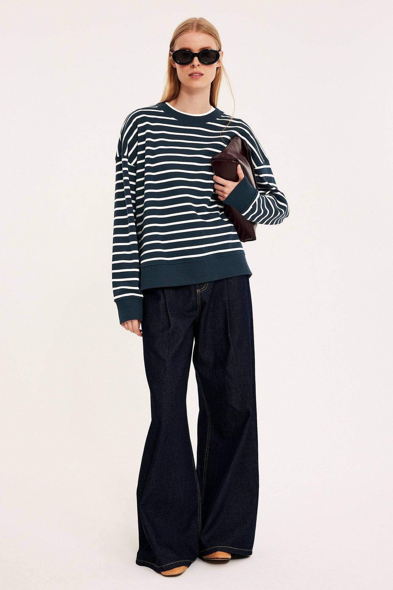 Forfra: Kvinne i mørkeblå stripet genser, vide mørkeblå bukser, svarte solbriller, holder brun clutch.