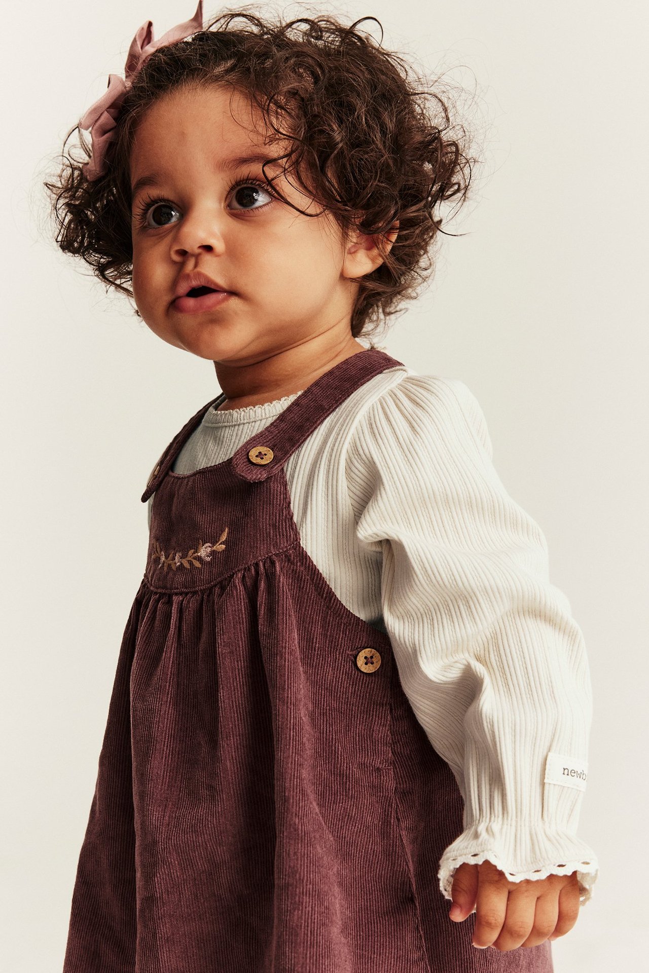 Frontvisning: baby i lys beige ribbet body med blondekanter, plommefarget kordfløyelkjole, rosa sløyfe.