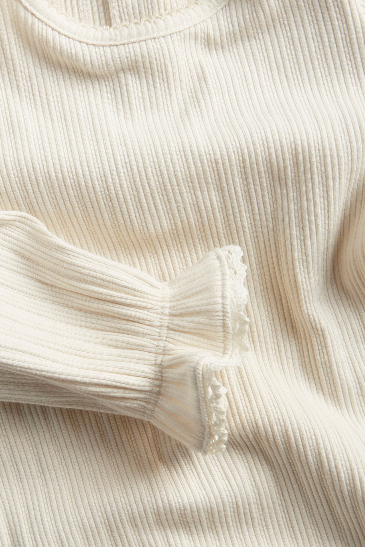 Zbliżenie na jasnobeżowe prążkowane body niemowlęce z falbanką, koronkowym mankietem i dekoltem z obszyciem picot.