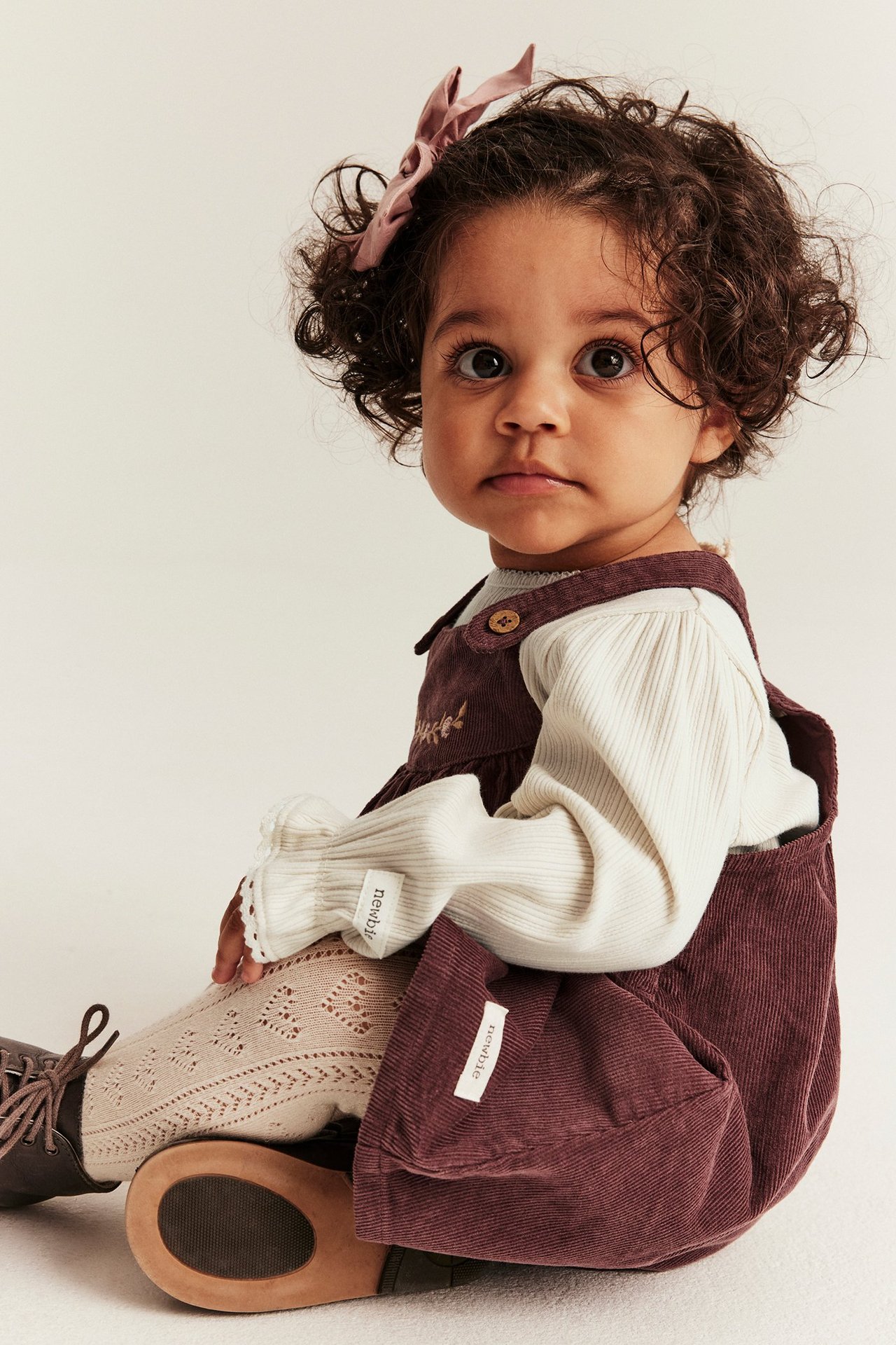 Baby i lys beige ribbet body med blondekanter, mørkerød kordfløyelssmekkebukse og mønstrede strømpebukser.