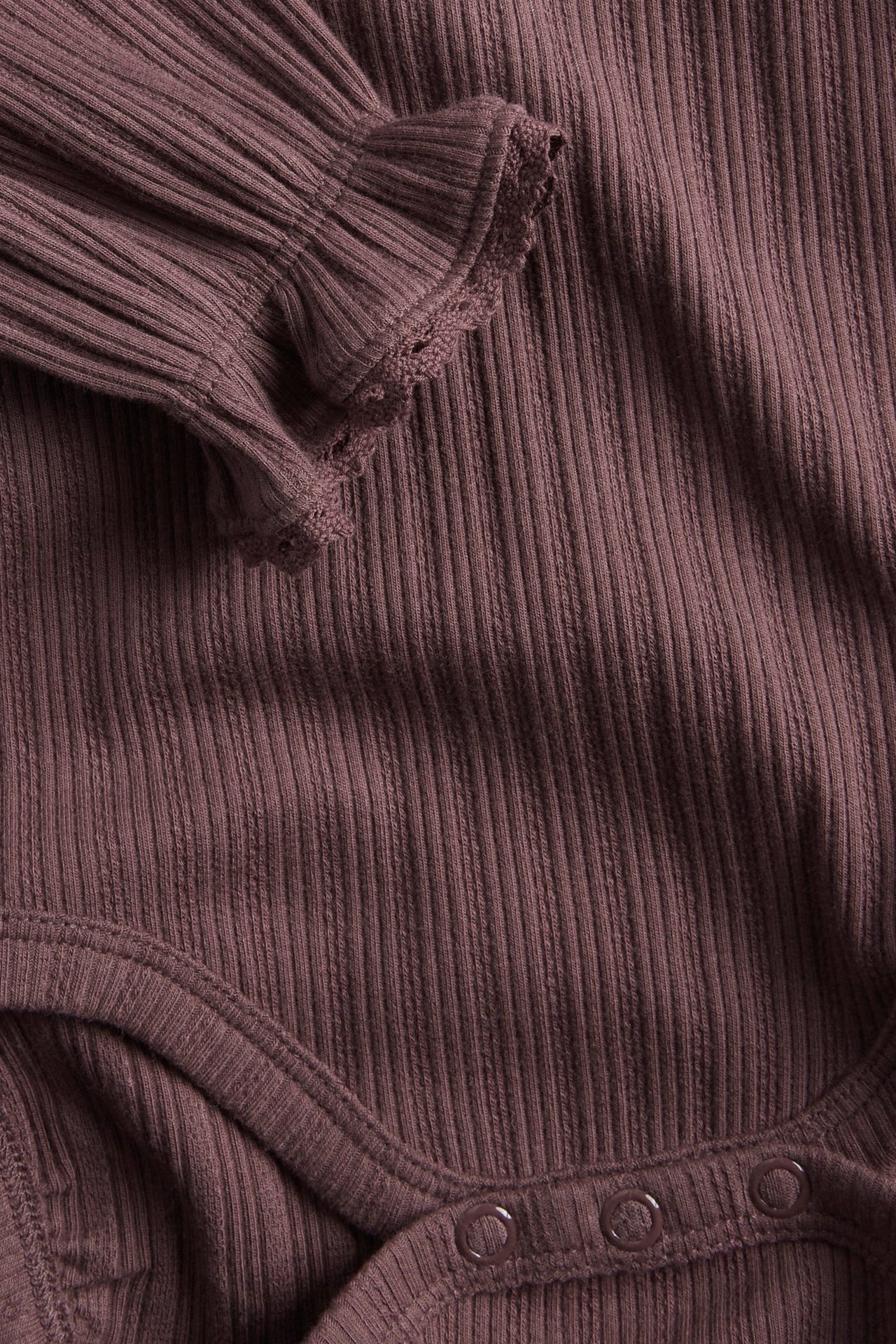 Närbild på en lila ribbad babybody med volang och spetskant vid ärmslut, tryckknappar.