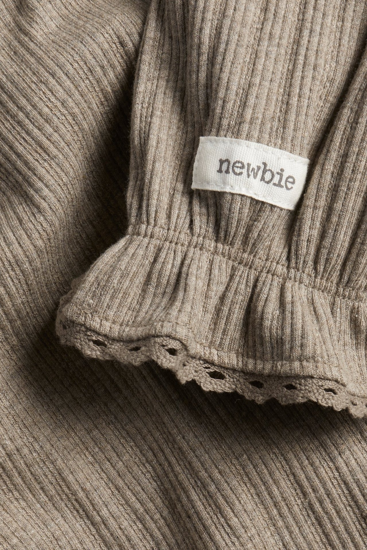 Lähikuva ruskeanmelange-värisestä ribbipaidasta, jossa on röyhelöinen, pitsireunainen hihansuu ja 'newbie'-merkki.