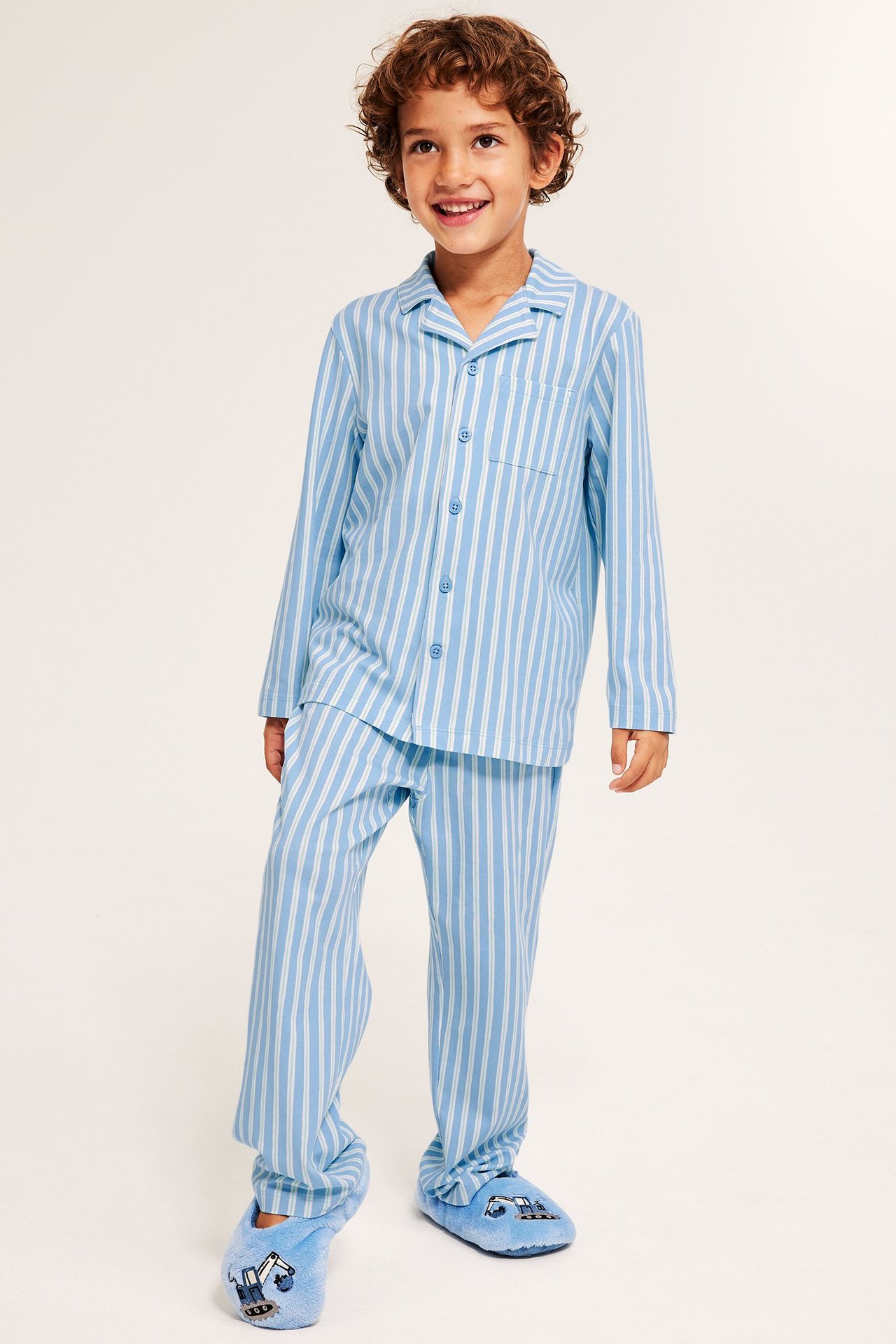 Widok z przodu: dziecko w jasnoniebieskiej piżamie w paski (zapinana góra, spodnie) i niebieskich kapciach-koparkach.