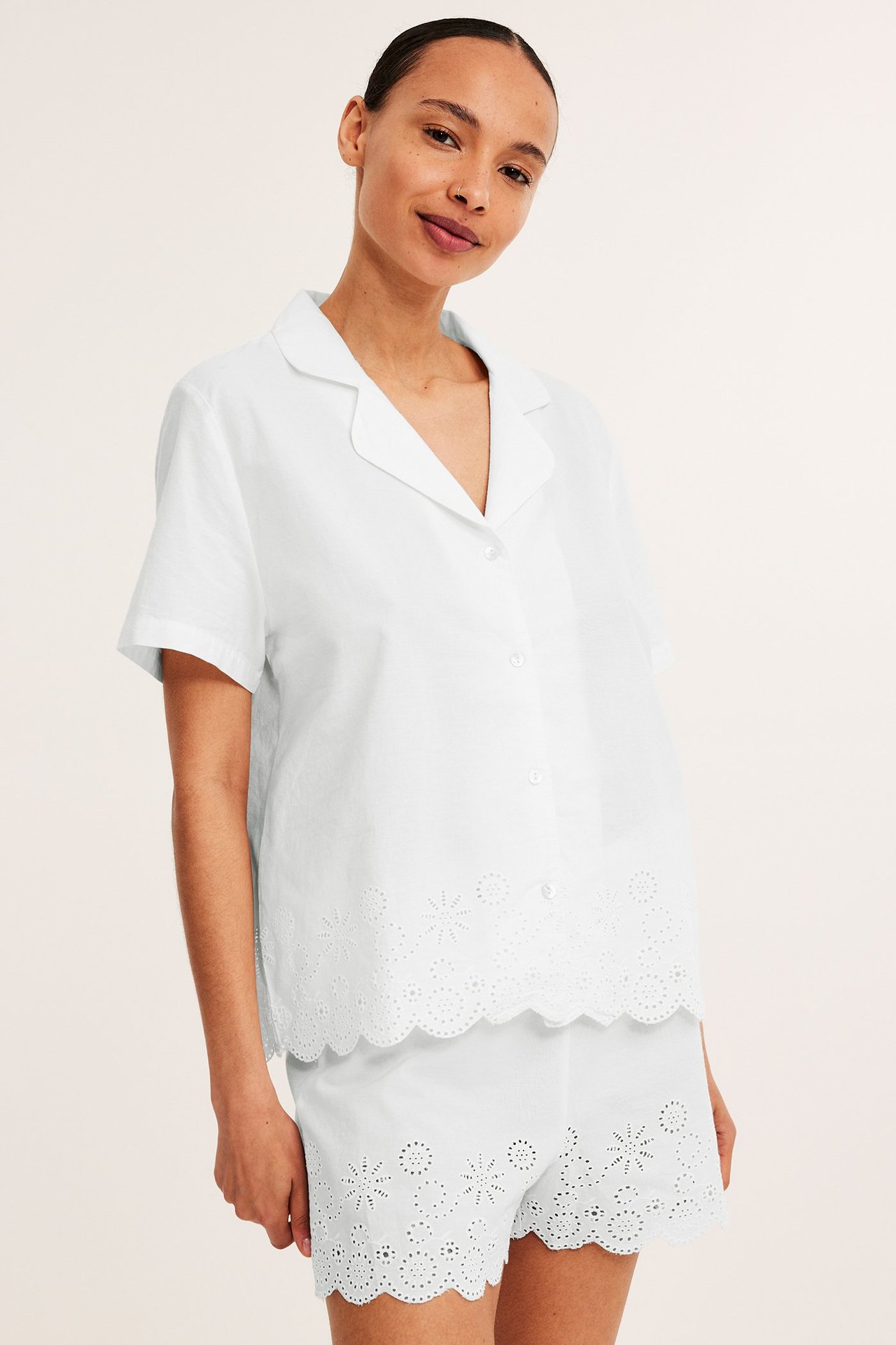 Edestä: nainen yllään luonnonvalkoinen puuvillapyjama, jossa lyhythihainen paita ja shortsit brodeeratuilla, aaltoilevilla reunoilla.