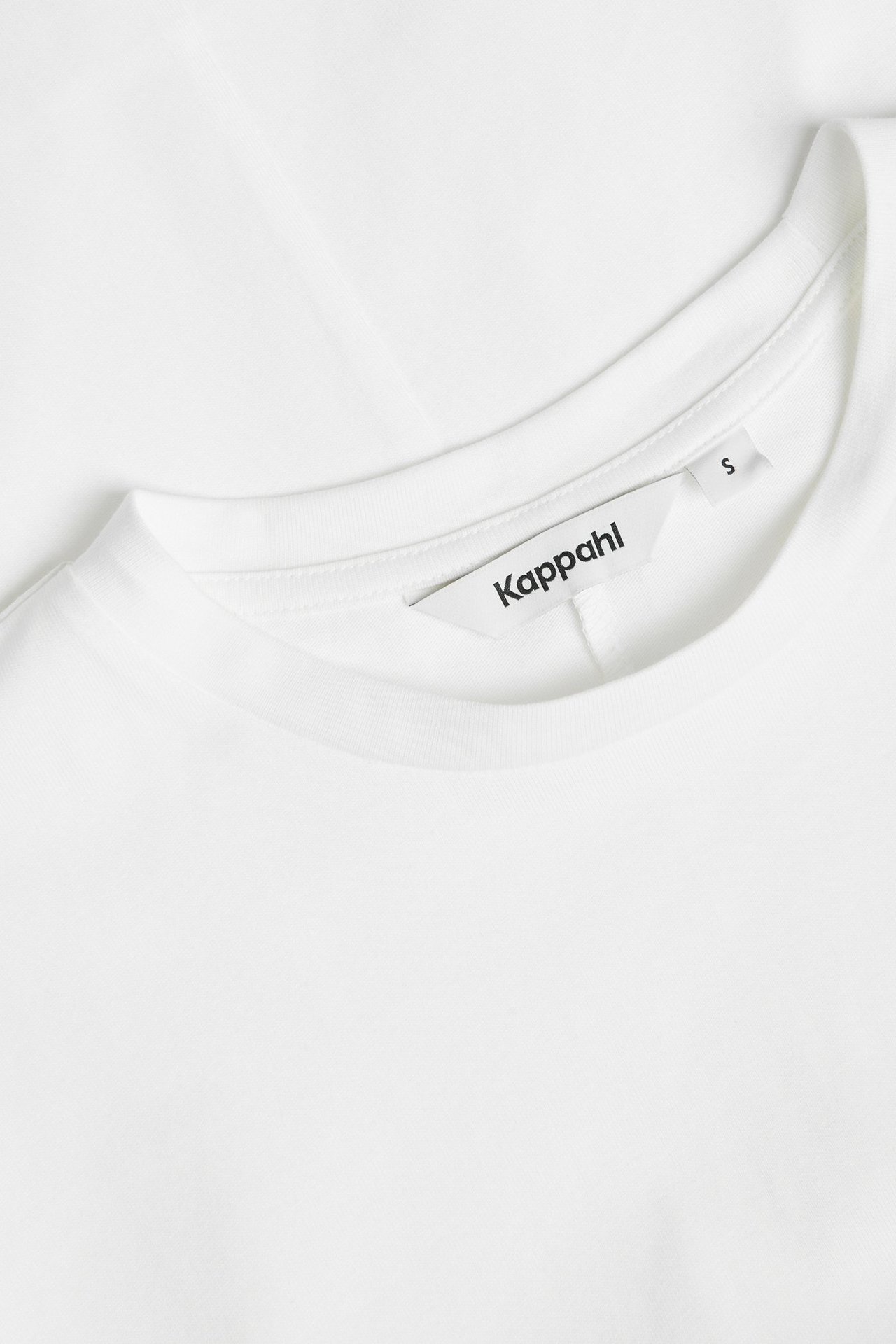 Zbliżenie dekoltu sukienki z bawełnianego dżerseju w kolorze złamanej bieli z okrągłym dekoltem i metką Kappahl.