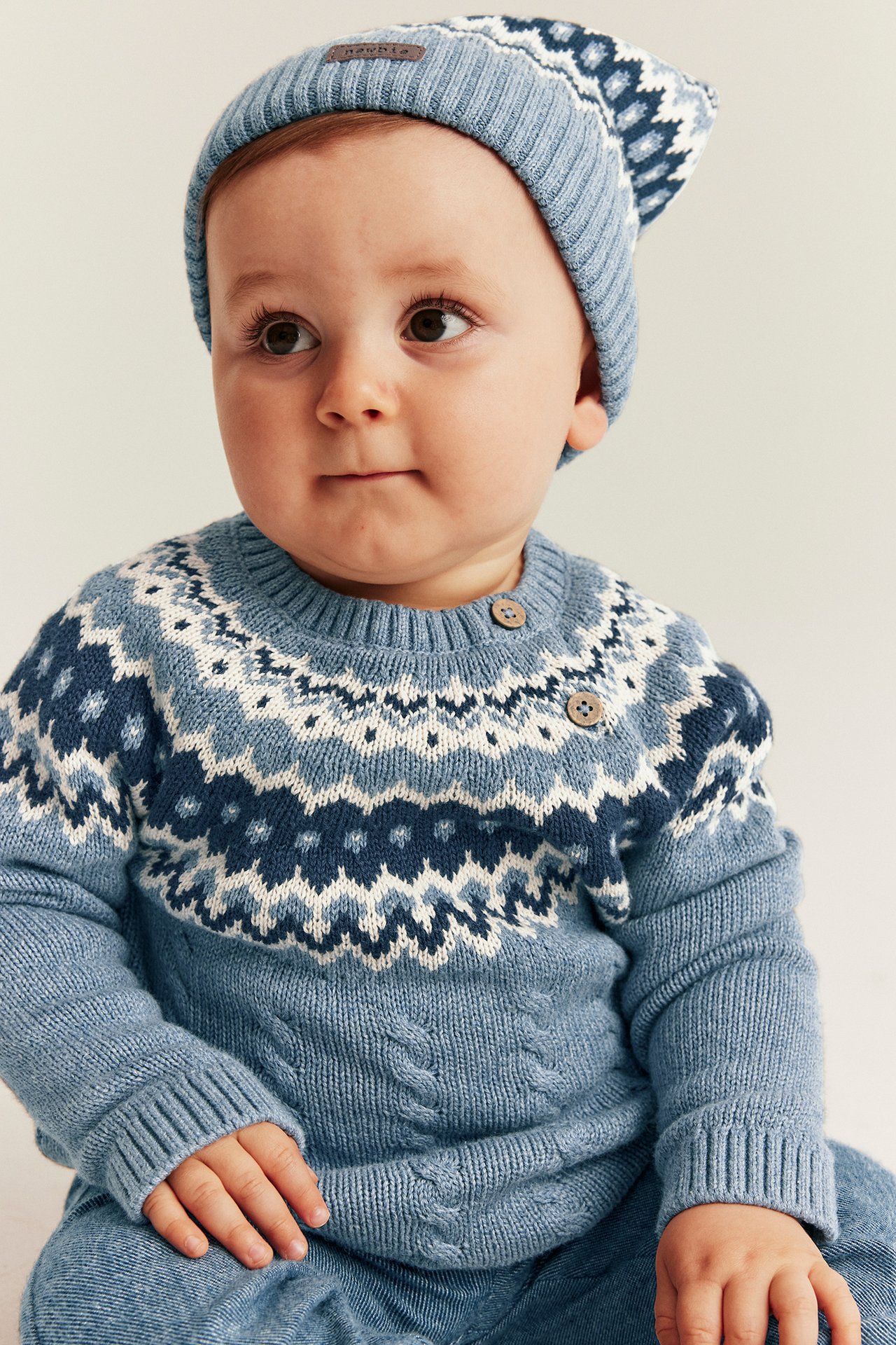 Nærbilde forfra av baby i blå jacquard- og flettestrikket genser med knapper, matchende lue og blå bukser.
