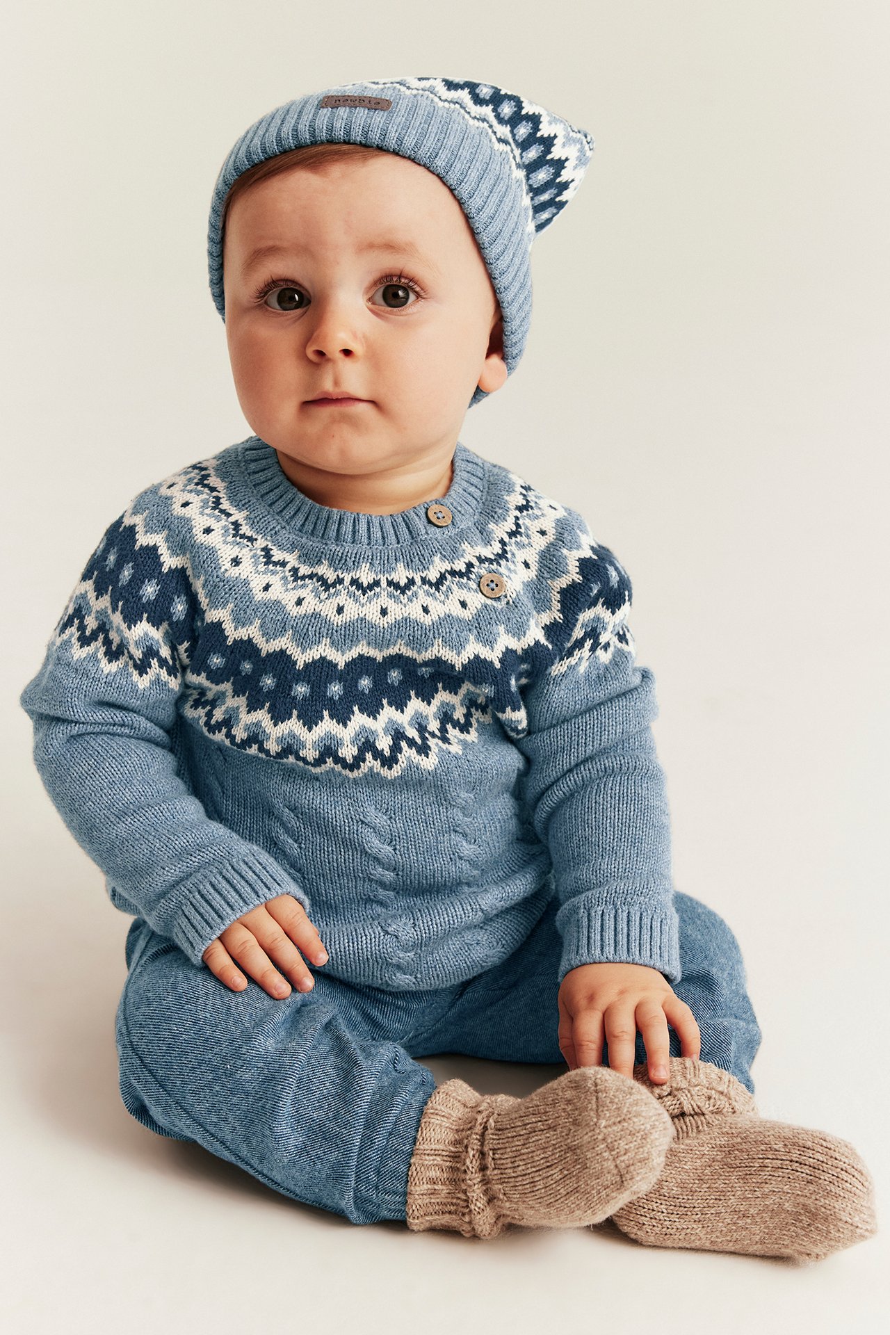 Frontbilde av en baby i blå jacquardstrikket genser, blå bukser, matchende lue og beige tøfler.