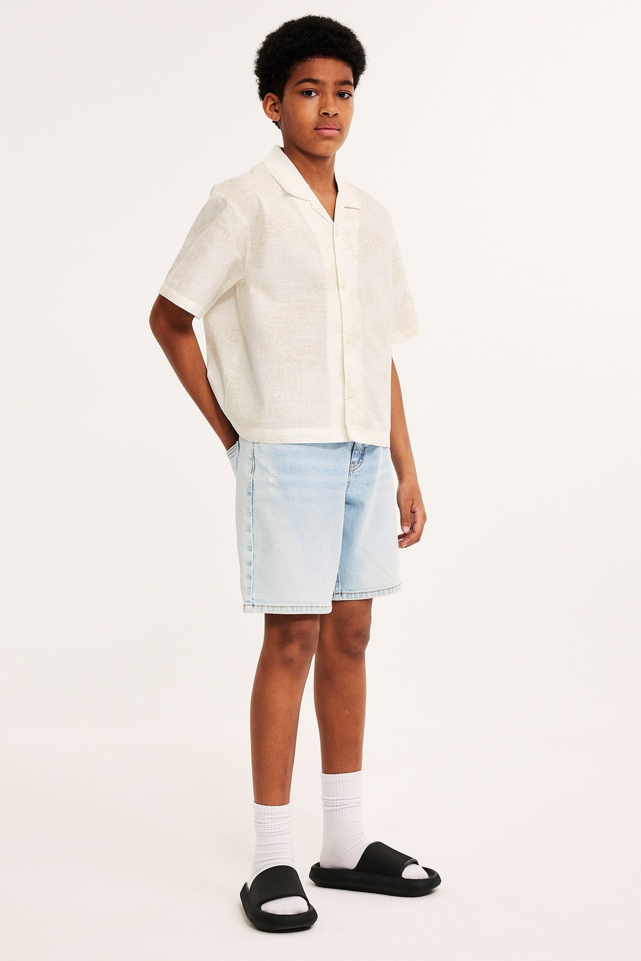 Widok z przodu: Chłopiec w kremowej, krótkiej koszuli bawełnianej w paisley, jasnoniebieskich szortach jeansowych, białych skarpetkach, czarnych klapkach.