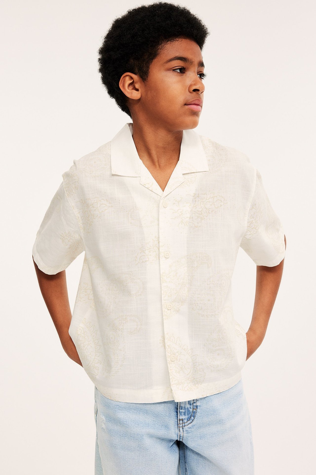 Widok z przodu: dziecko w kremowej, krótkiej koszuli bawełnianej z paisley i jasnoniebieskich dżinsach.