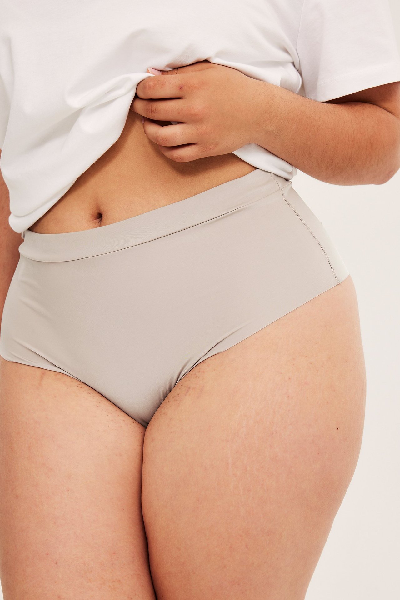 Lähikuva edestä naisesta, jolla on vaaleanpunaiset korkeavyötäröiset mikroalushousut ja valkoinen t-paita.