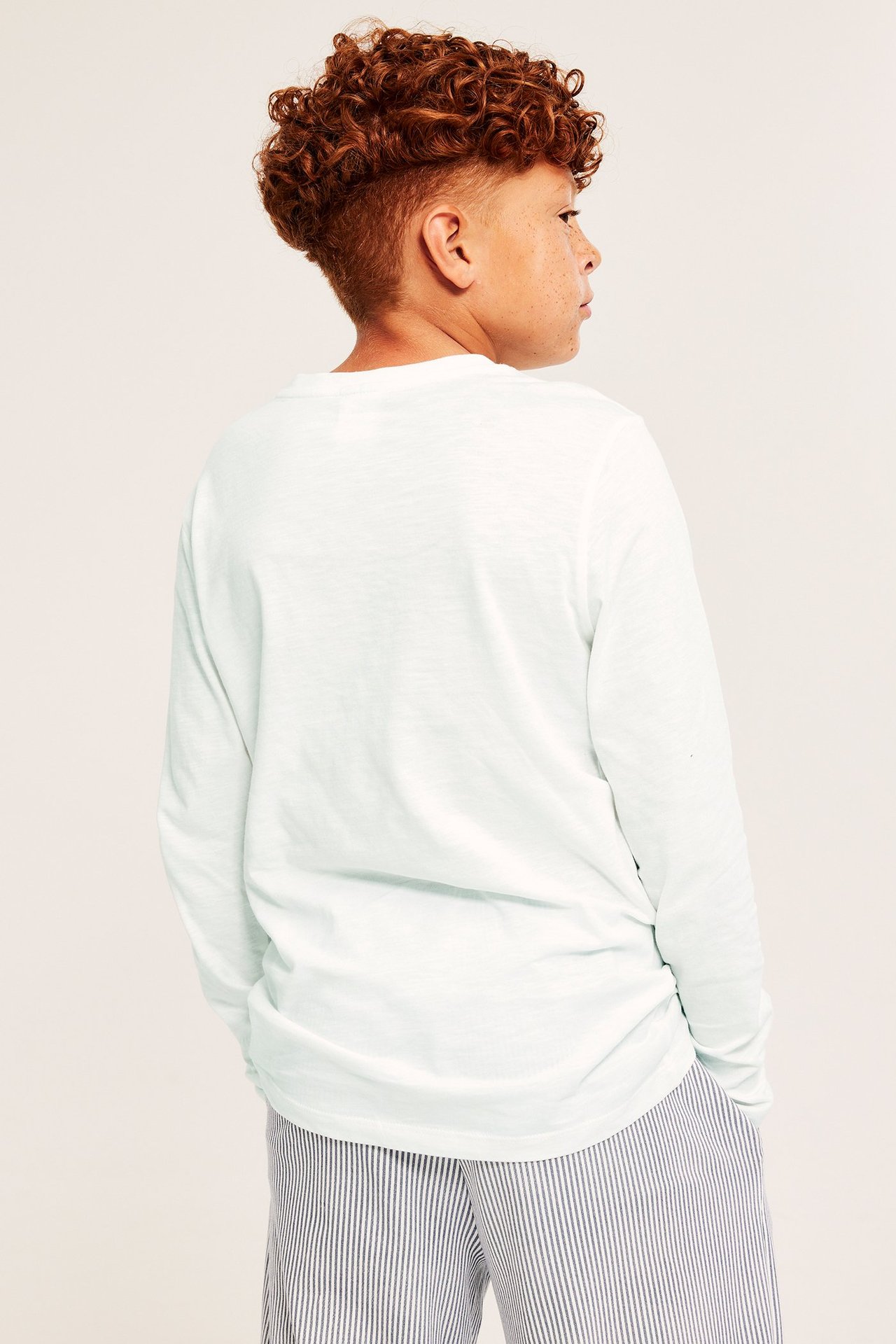 Barn, bakifrån, i en offwhite långärmad bomulls-t-shirt och blåvitrandiga byxor.