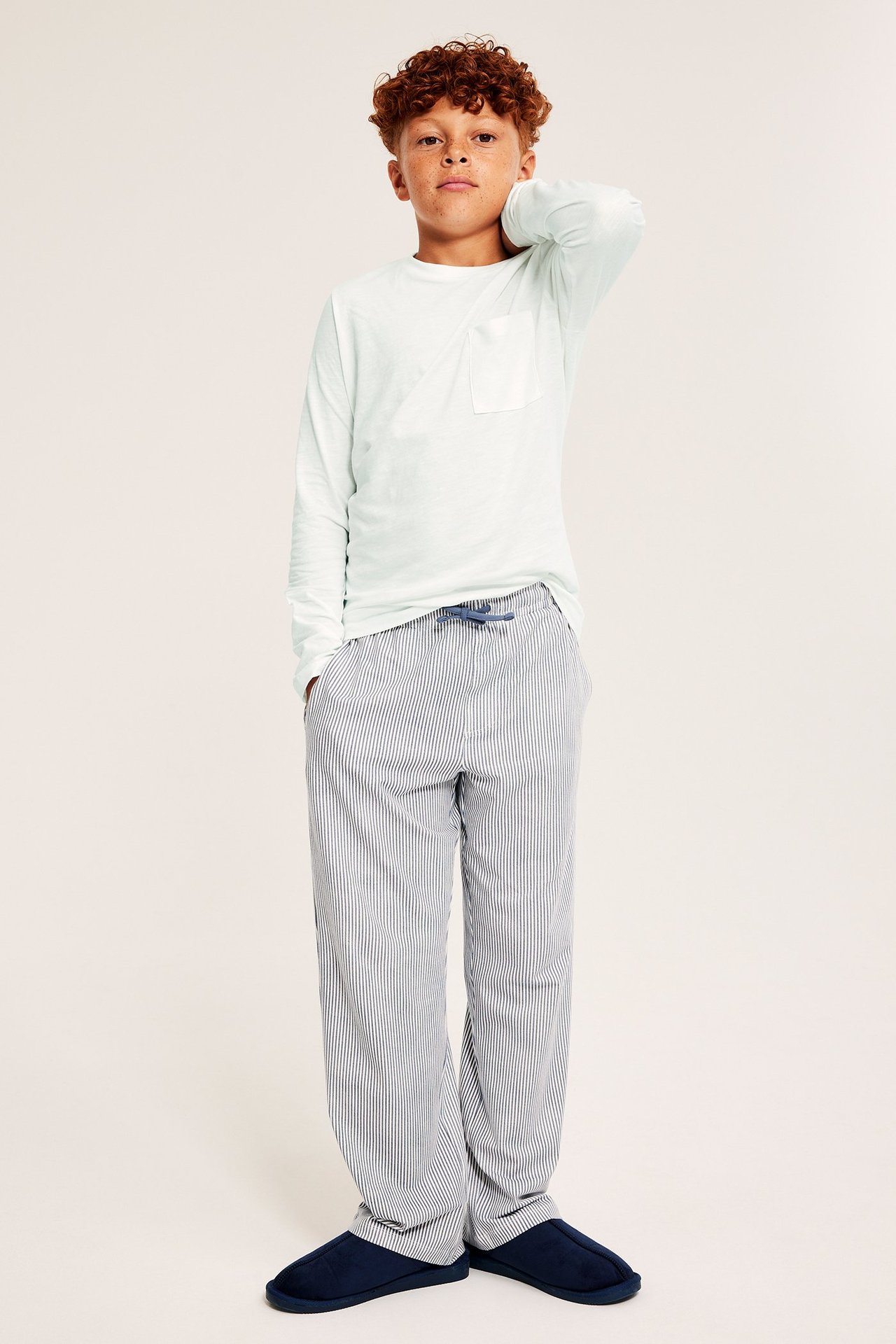 Frontvisning: En pojke i en offwhite långärmad slub-t-shirt med ficka, blårandiga byxor och mörkblå tofflor.