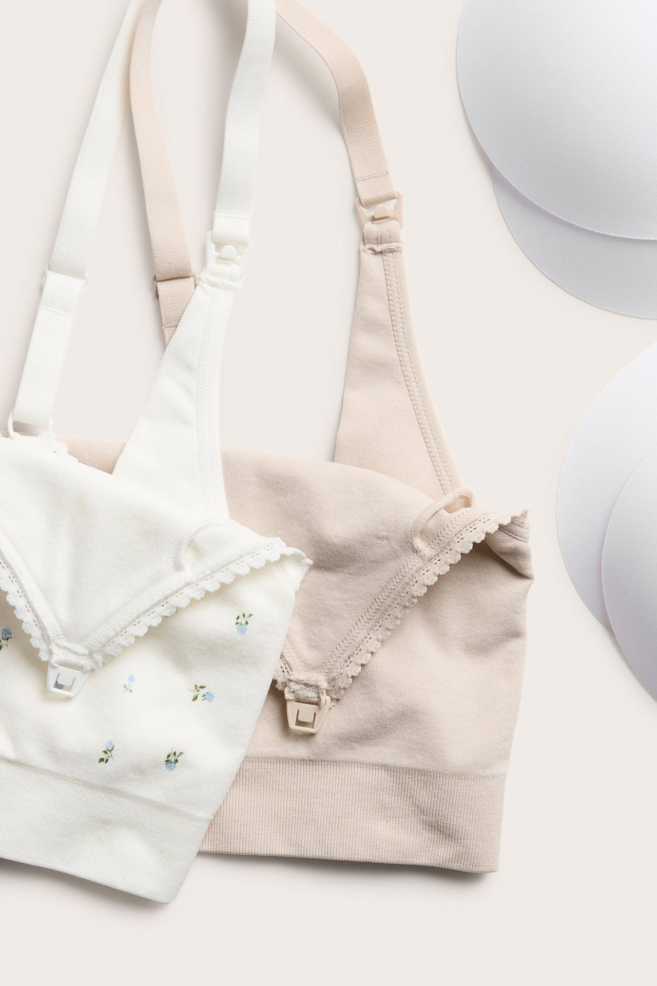 Zbliżenie dwóch biustonoszy do karmienia: jeden ecru w kwiaty, drugi beżowy, z dwoma białymi wkładkami.