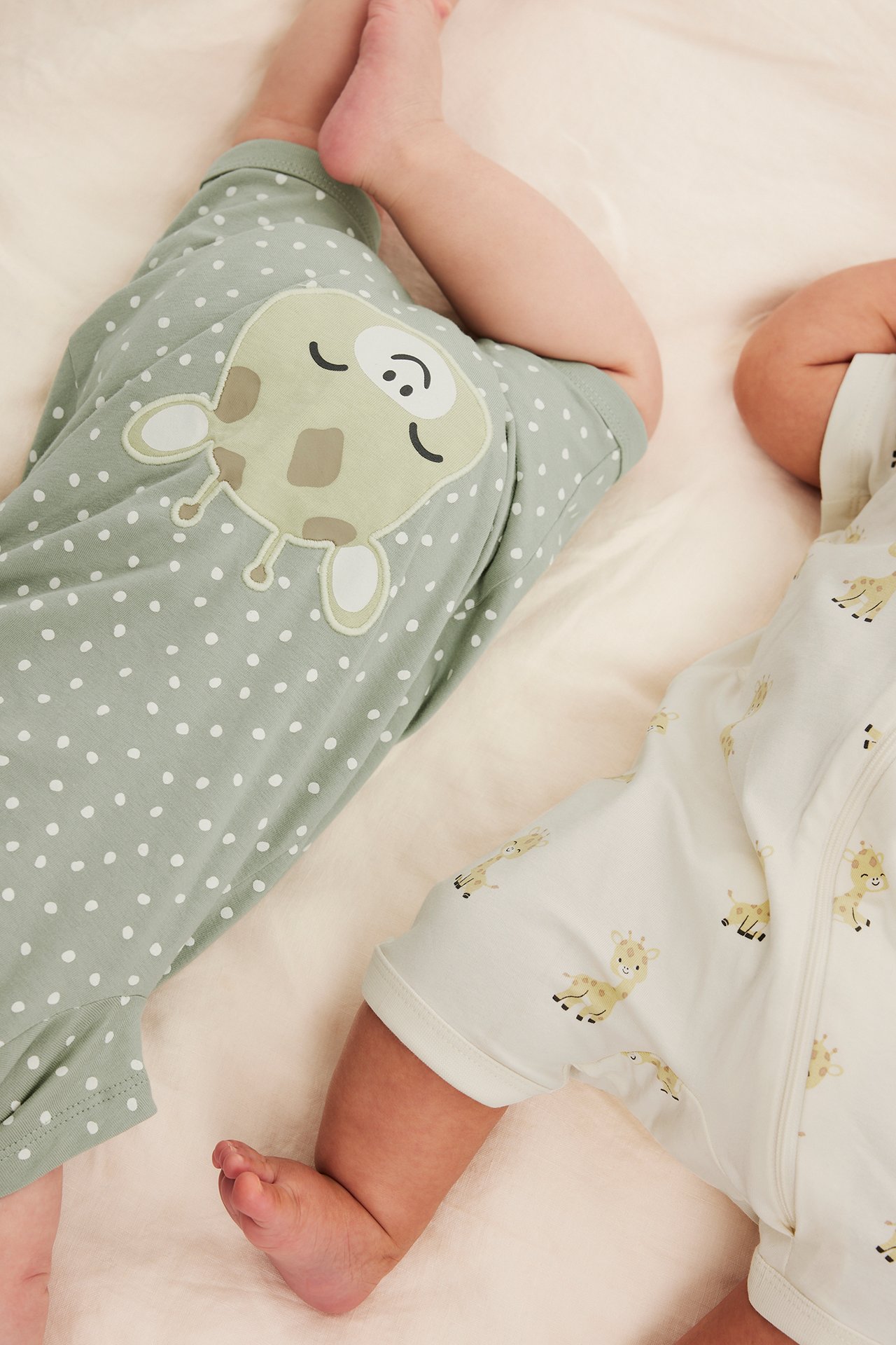 Toppvy av två bebisar i kortärmade rompers: en offwhite med girafftryck, en grön med prickar och giraffapplikation.