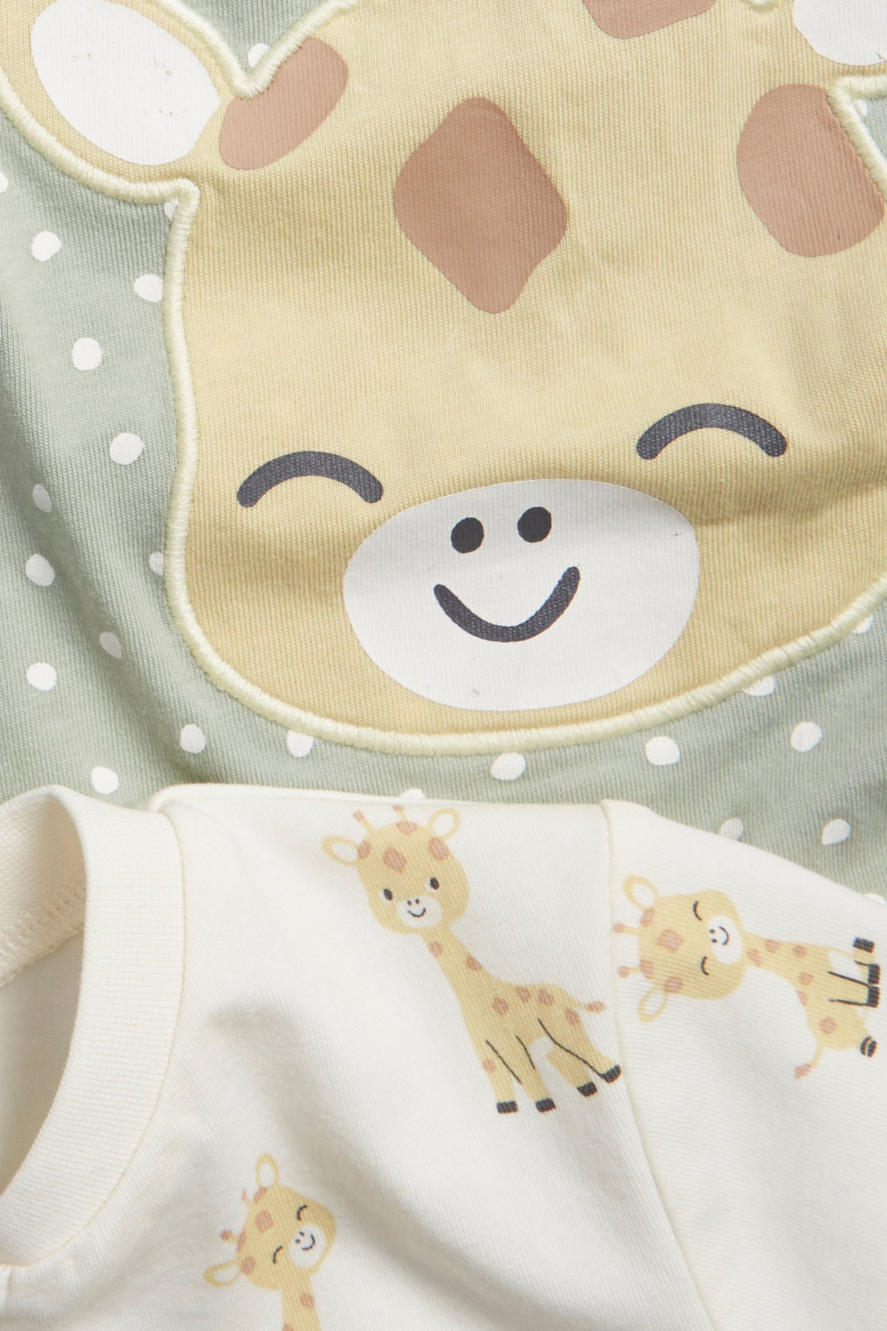 Närbild på två kortärmade babyrompers: en ljusgrön med giraffansikte-applikation, en offwhite med giraffmönster.