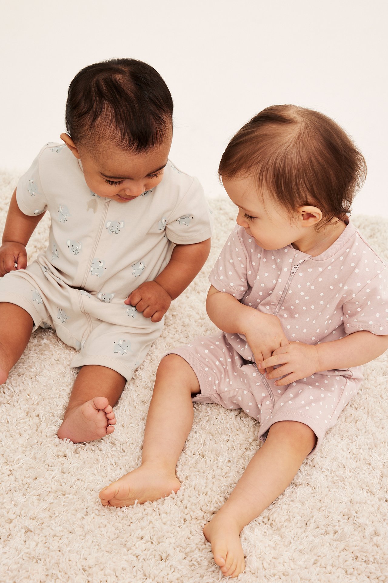 Kaksi vauvaa lyhythihaisissa romppereissa: toinen beige norsukuvioinen, toinen vaaleanpunainen pilkullinen.