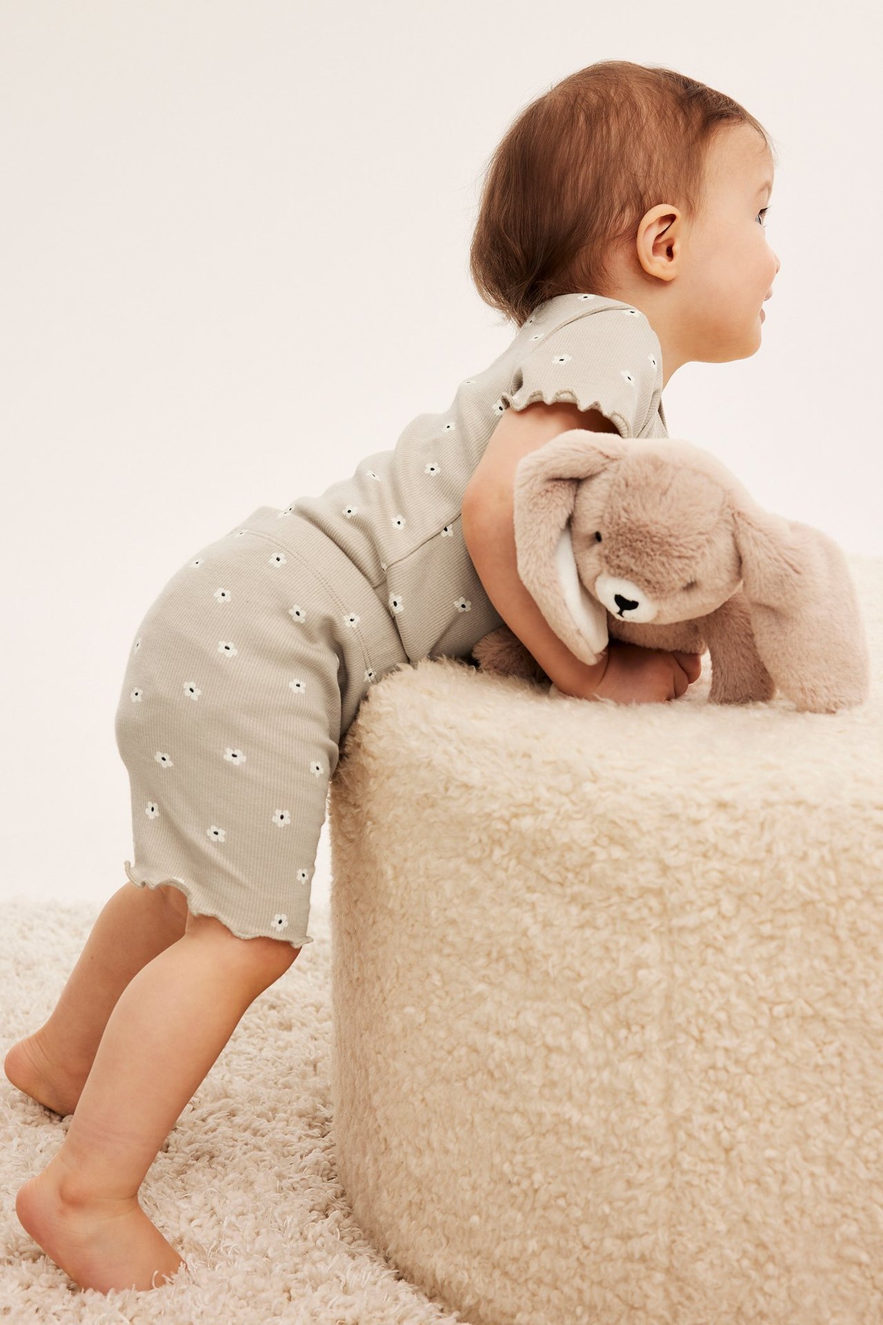 Sivukuva vauvasta ribatussa vaaleanbeigessä kukkakuosisessa romperissa pyöräilyshortsein, pitäen pehmopupua.