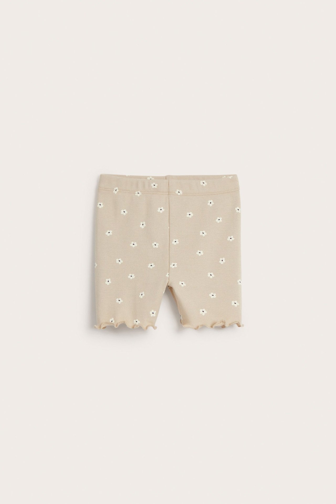 Edestä näkymä beigestä ribbineulotusta vauvan pyöräilyshortsista, jossa kukkakuvio ja röyhelöreunat.