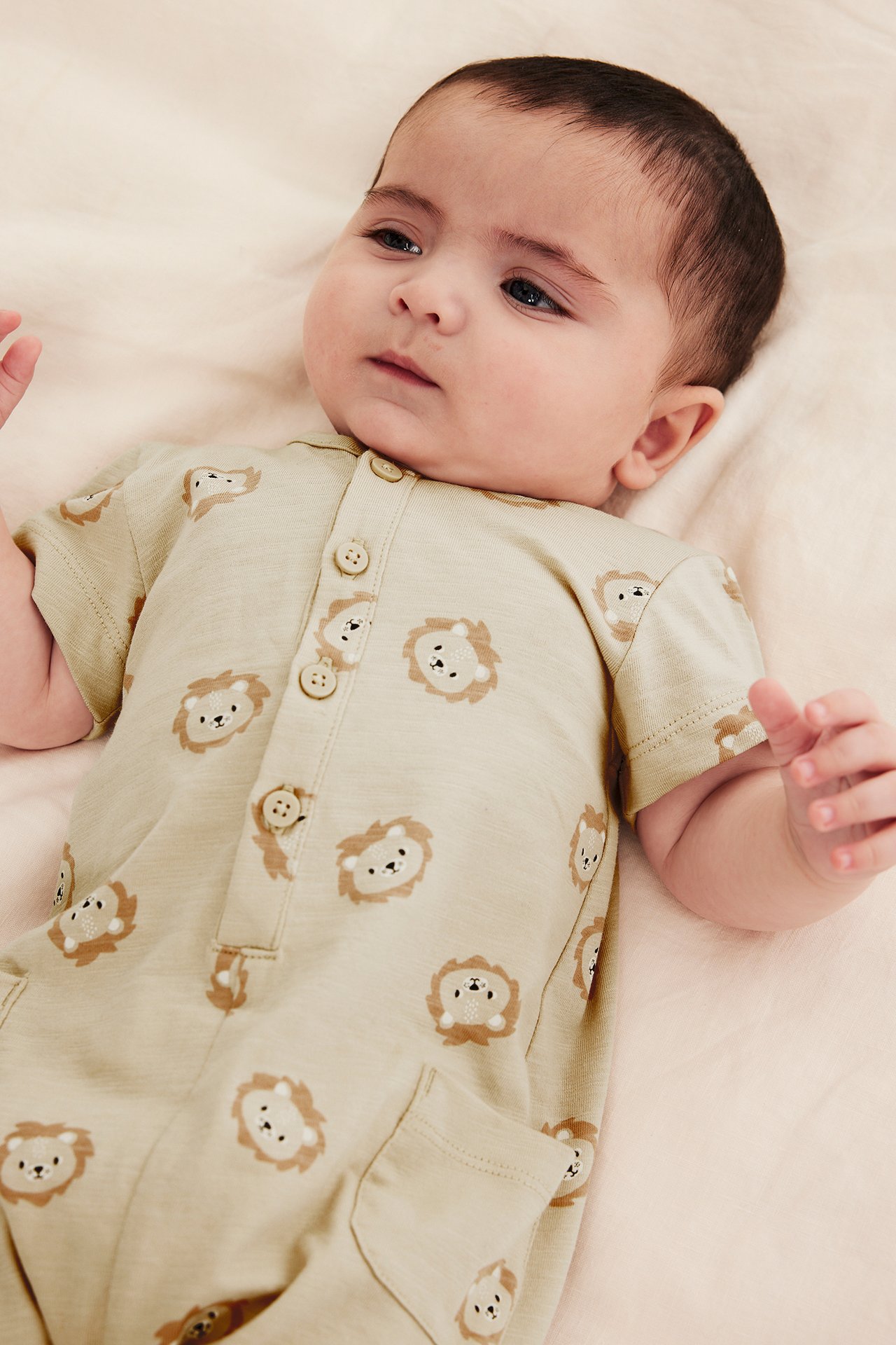 Nærbilde av baby i beige kortermet bomullsjumpsuit med løvemønster og knapper foran.