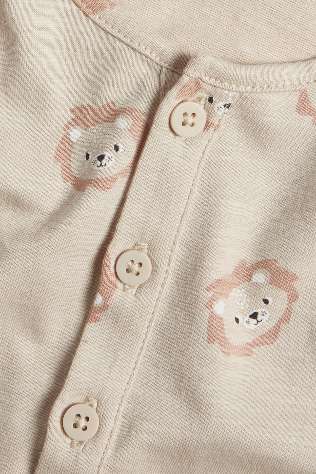 Nærbilde av en beige bomulls-babyjumpsuit med løvemønster og knapper foran.