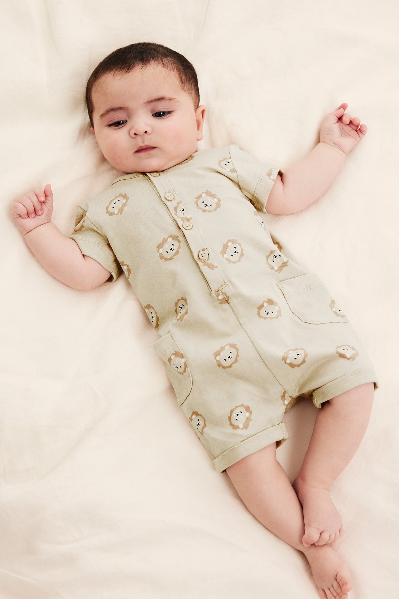 Ovenfra-visning av en baby i en beige kortermet playsuit med løveansiktsmønster, knapper foran og lommer.