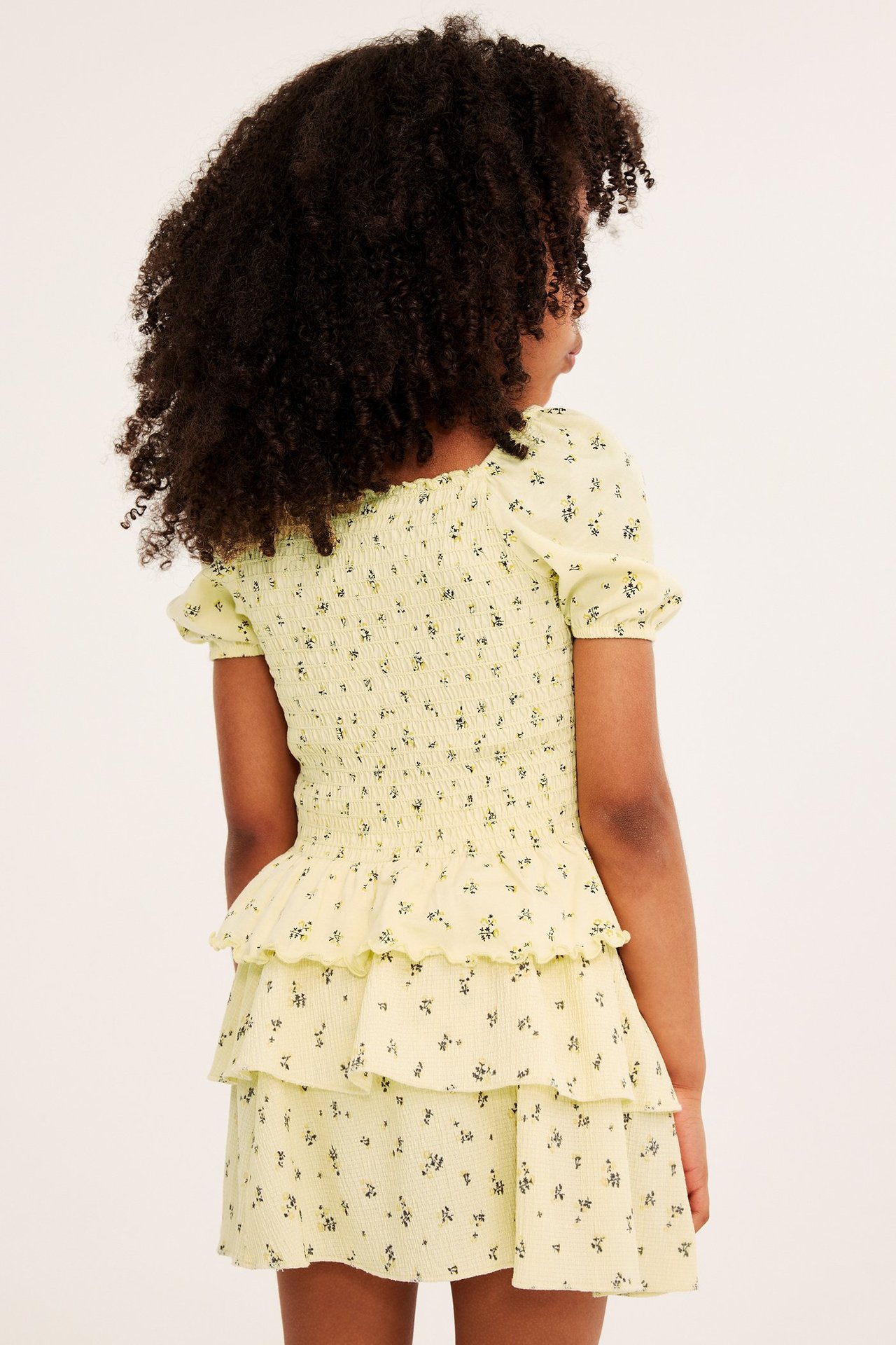 Bakifrån på ett barn i ljusbeige blommig smockad topp med puffärm och matchande volangkjol.