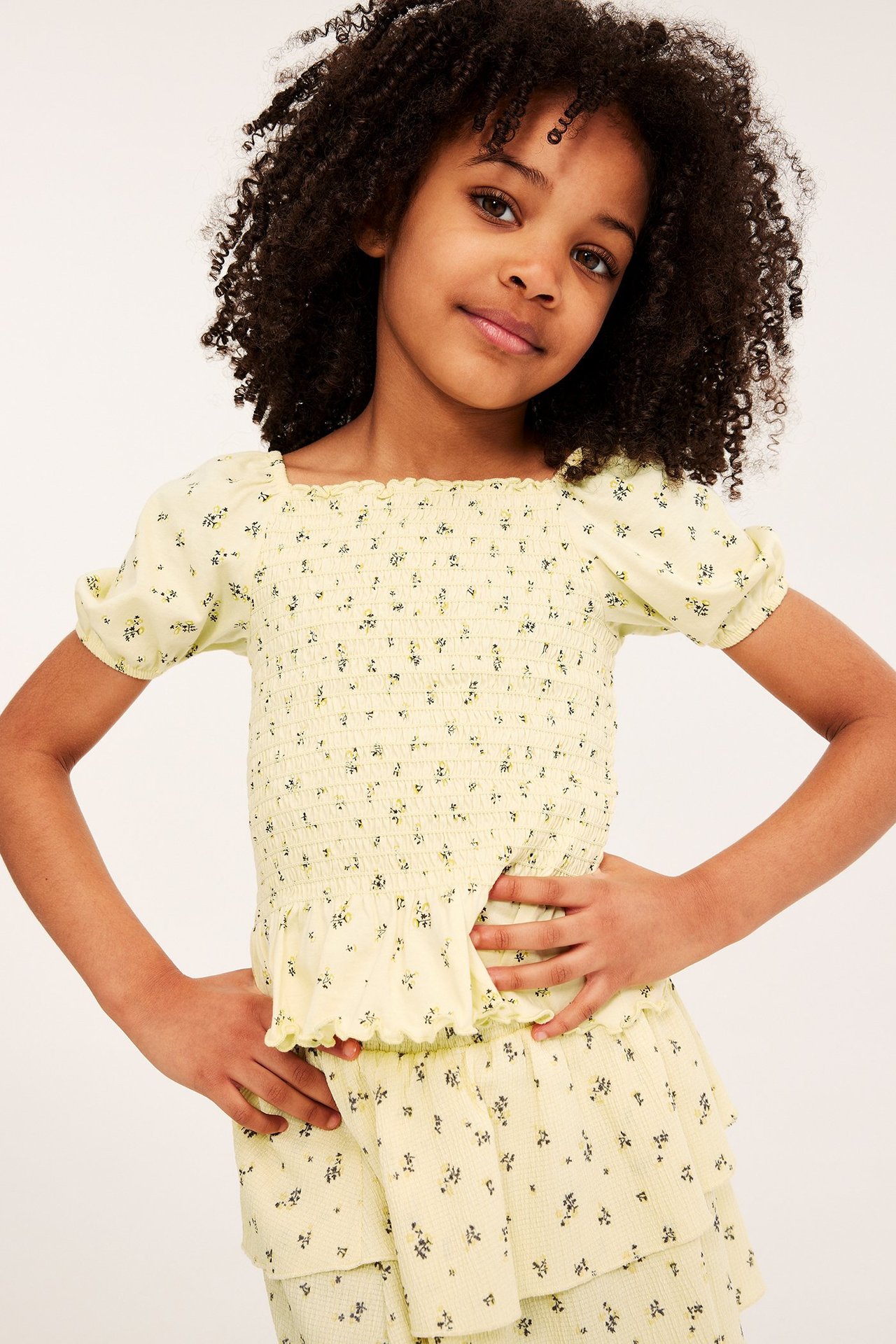 Framsida av en tjej som bär en ljusgul smockad blommig topp med korta puffärmar och matchande volangkjol.