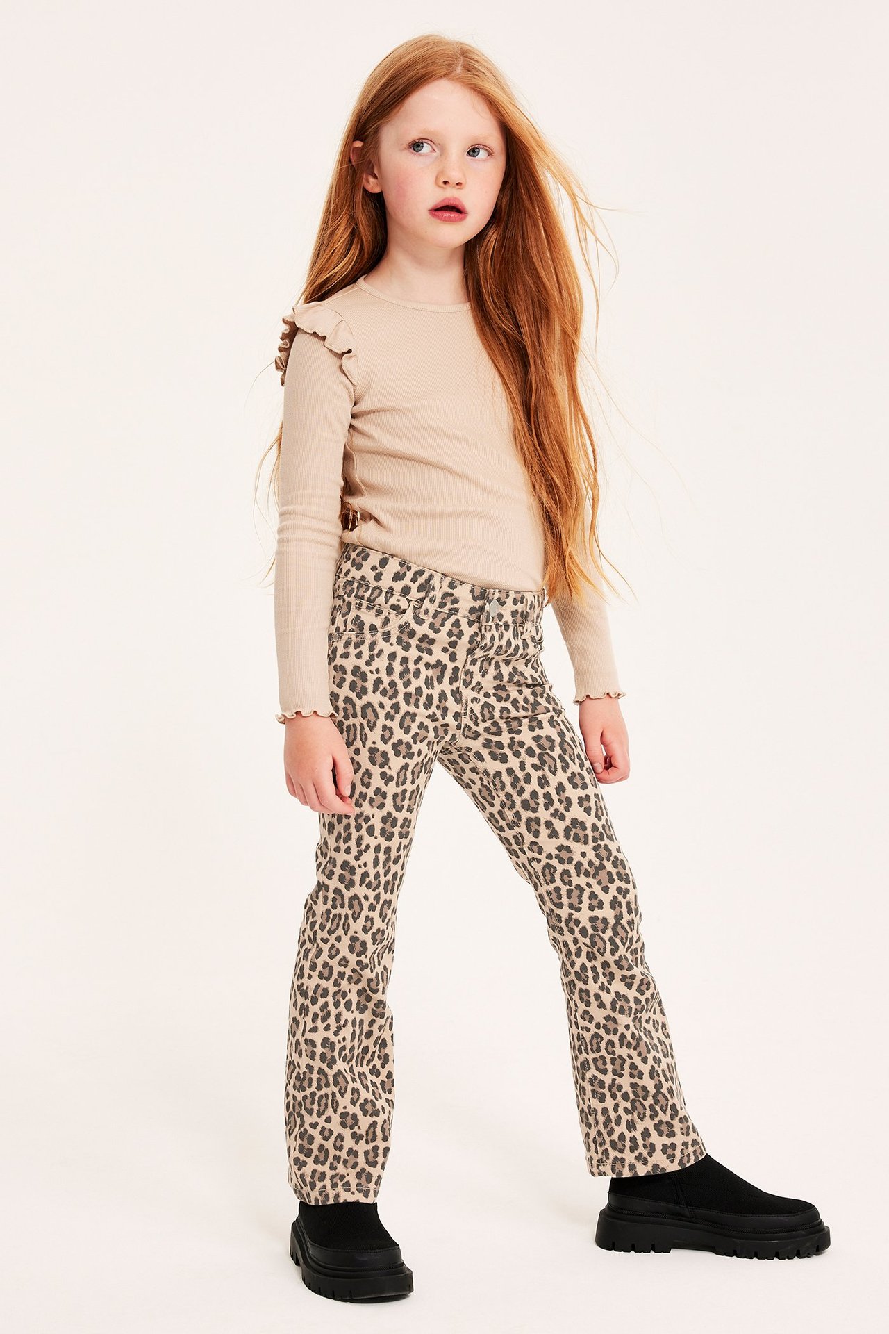Widok z przodu: dziecko w beżowych jeansach bootcut w panterkę, beżowej bluzce z długim rękawem i czarnych butach.