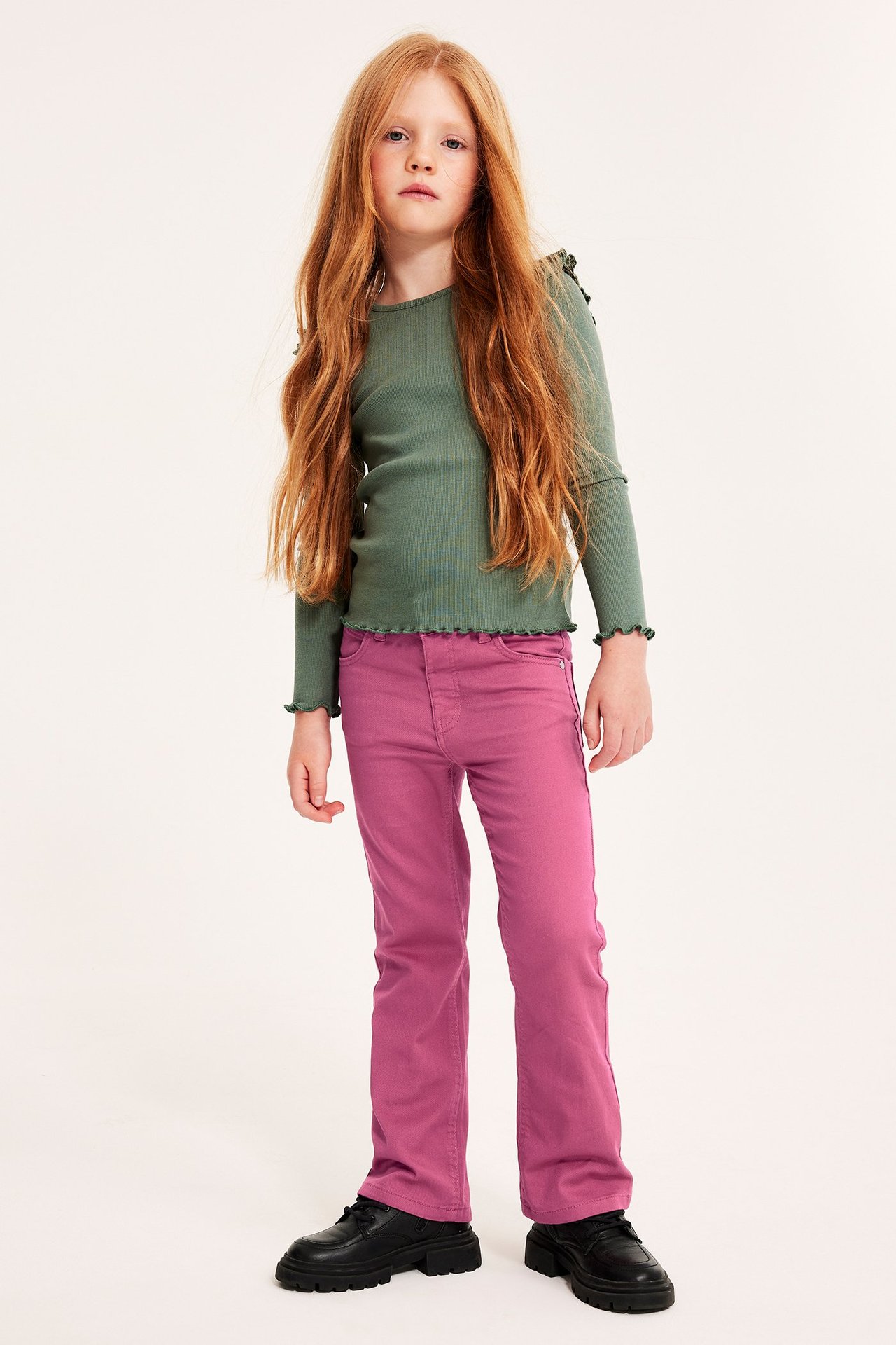 Widok z przodu dziecka w ciemnoróżowych jeansach bootcut, zielonej bluzce z długim rękawem i czarnych butach.