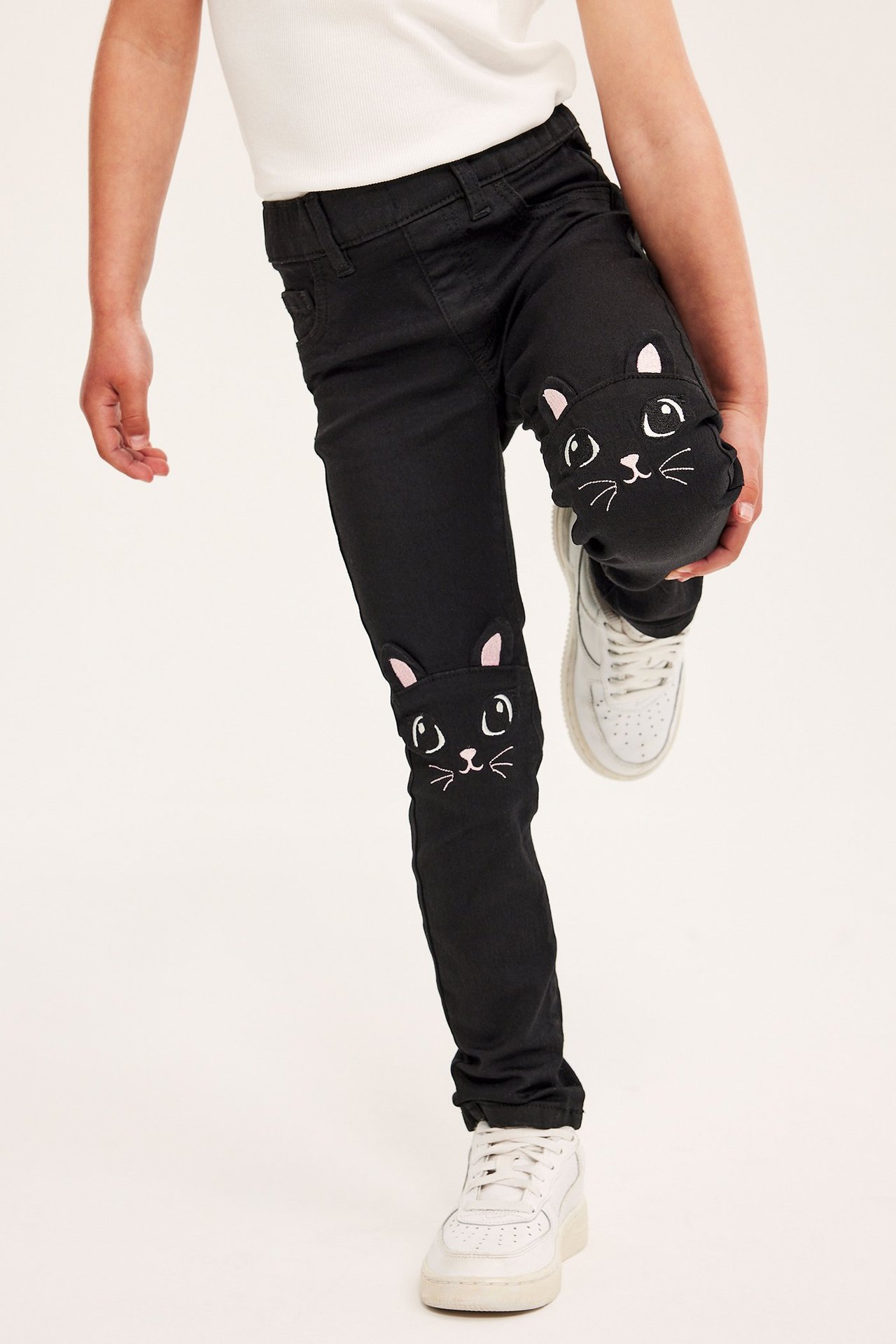 Edestä: Lapsi yllään mustat kapeat stretch-denim-jeggingit kissakirjonnalla polvissa, valkoinen t-paita, valkoiset lenkkarit.