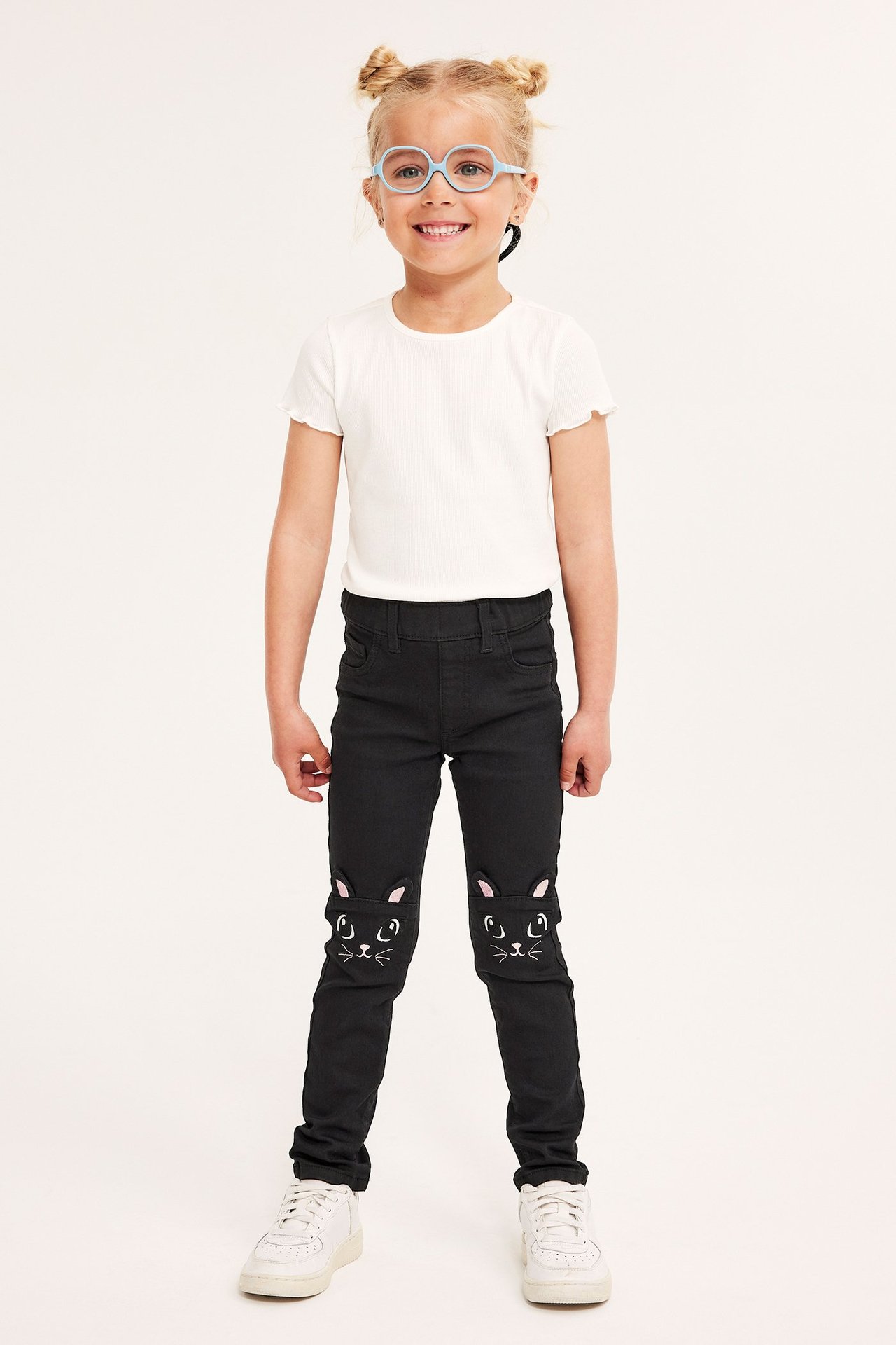 Przód: Dziecko w czarnych jegginsach z haftem kota, białym t-shircie, niebieskich okularach, białych butach.