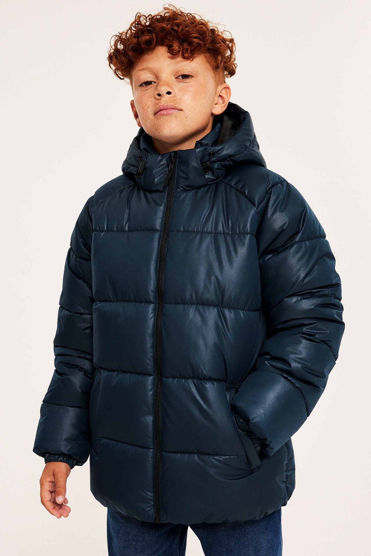 Framifrån, ett barn i mörkblå halvblank pufferjacka med huva och dragkedja, samt blå jeans.