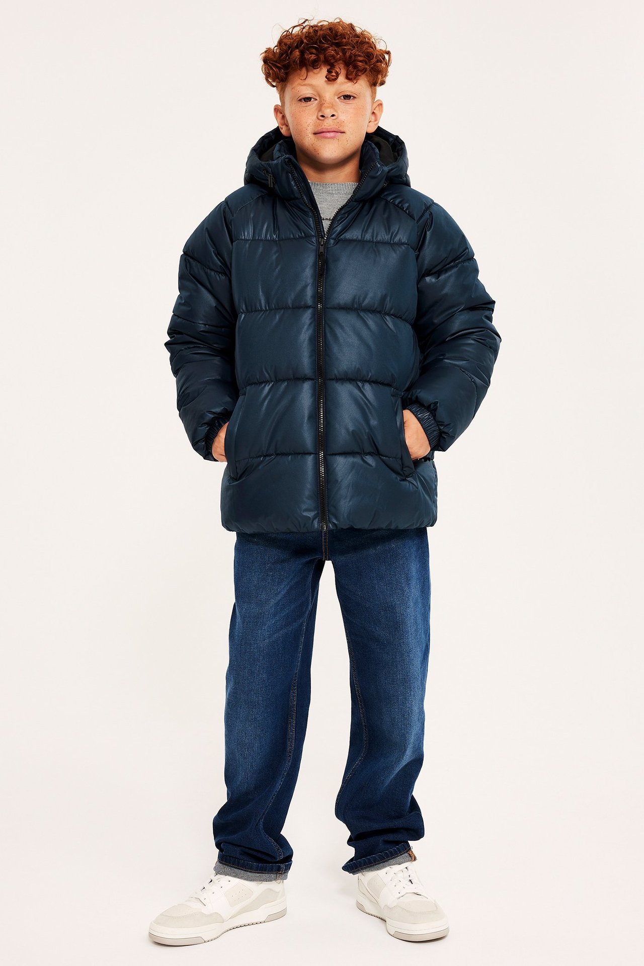 Hel frontvisning av et barn i en mørkeblå halvblank pufferjakke med hette, blå jeans og hvite joggesko.