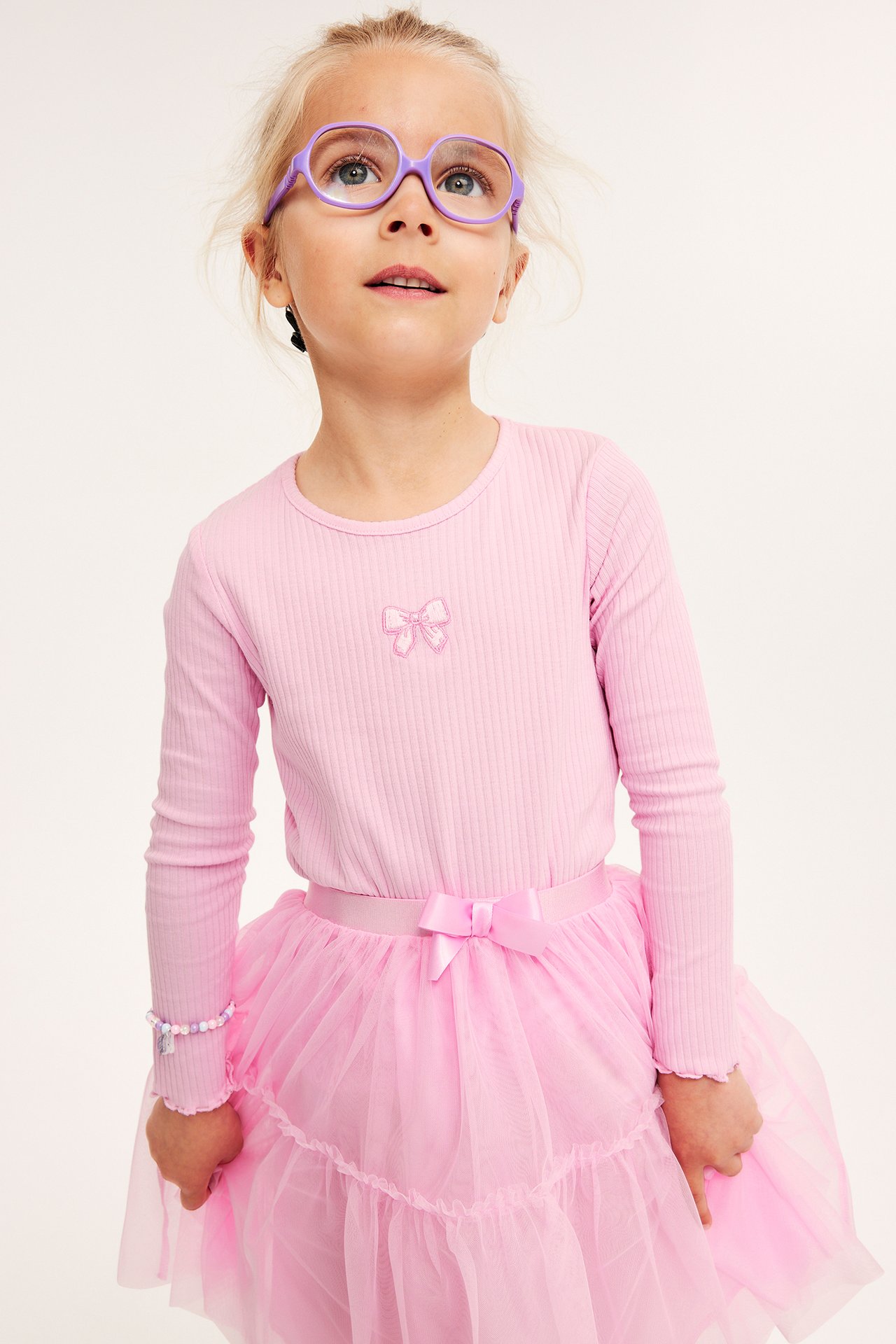Framsida: Barn i rosa ribbad långärmad topp med rosett, rosa tyllkjol, lila glasögon och armband.