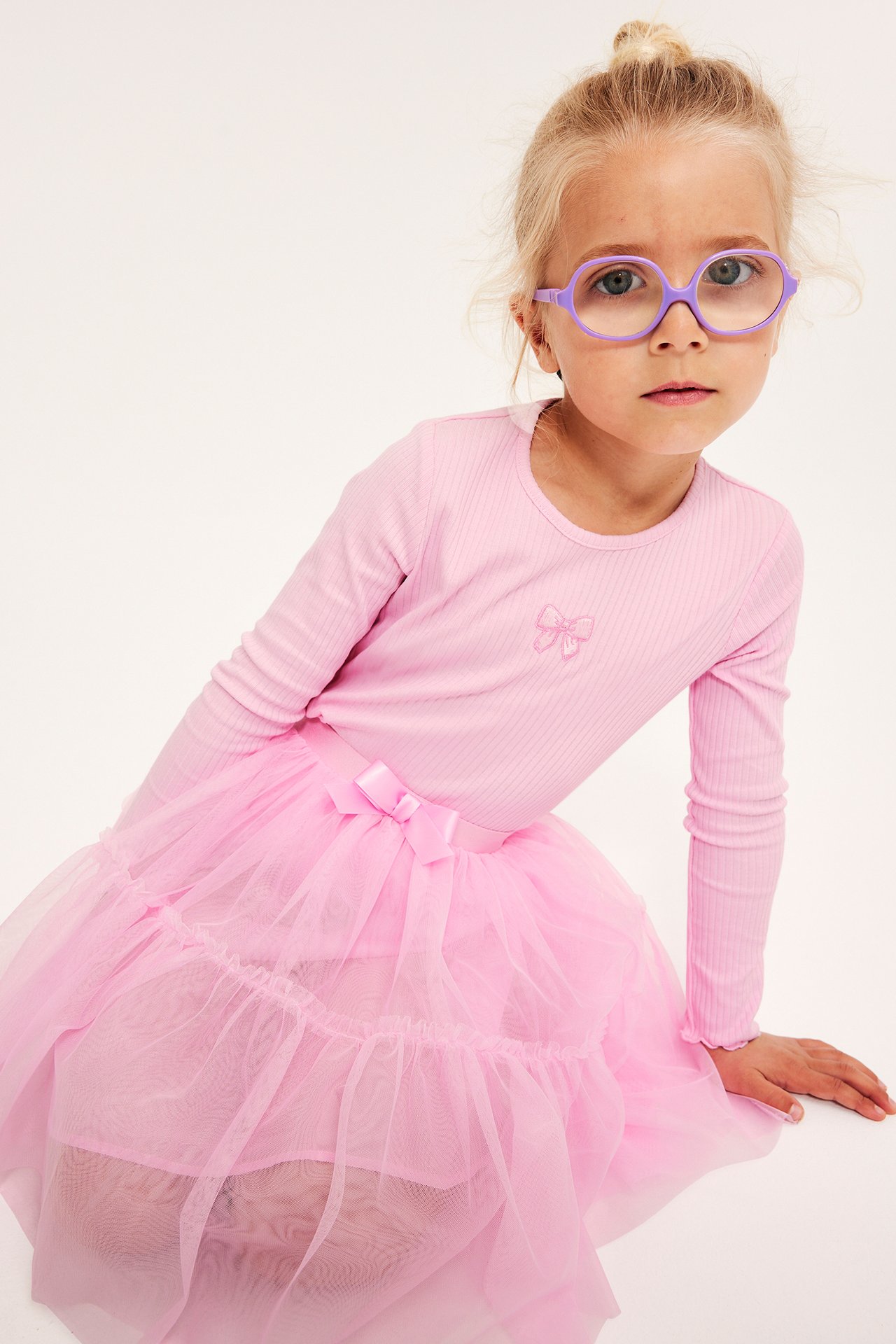 Barn i rosa ribbad långärmad topp med broderad rosett, rosa tyllkjol och lila glasögon.